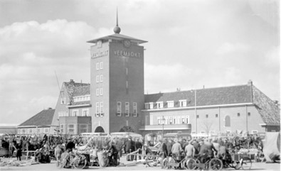 Black and white photograph of a crowded livestock market in front of the Vee Markt building, featuring a tall brick tower with a spire and people gathered around carts and bicycles