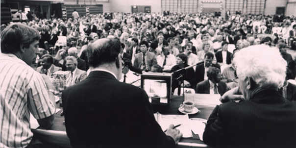 Black and white photograph showing a crowded indoor auction or meeting, with three men seated at a front table facing a large audience of people.