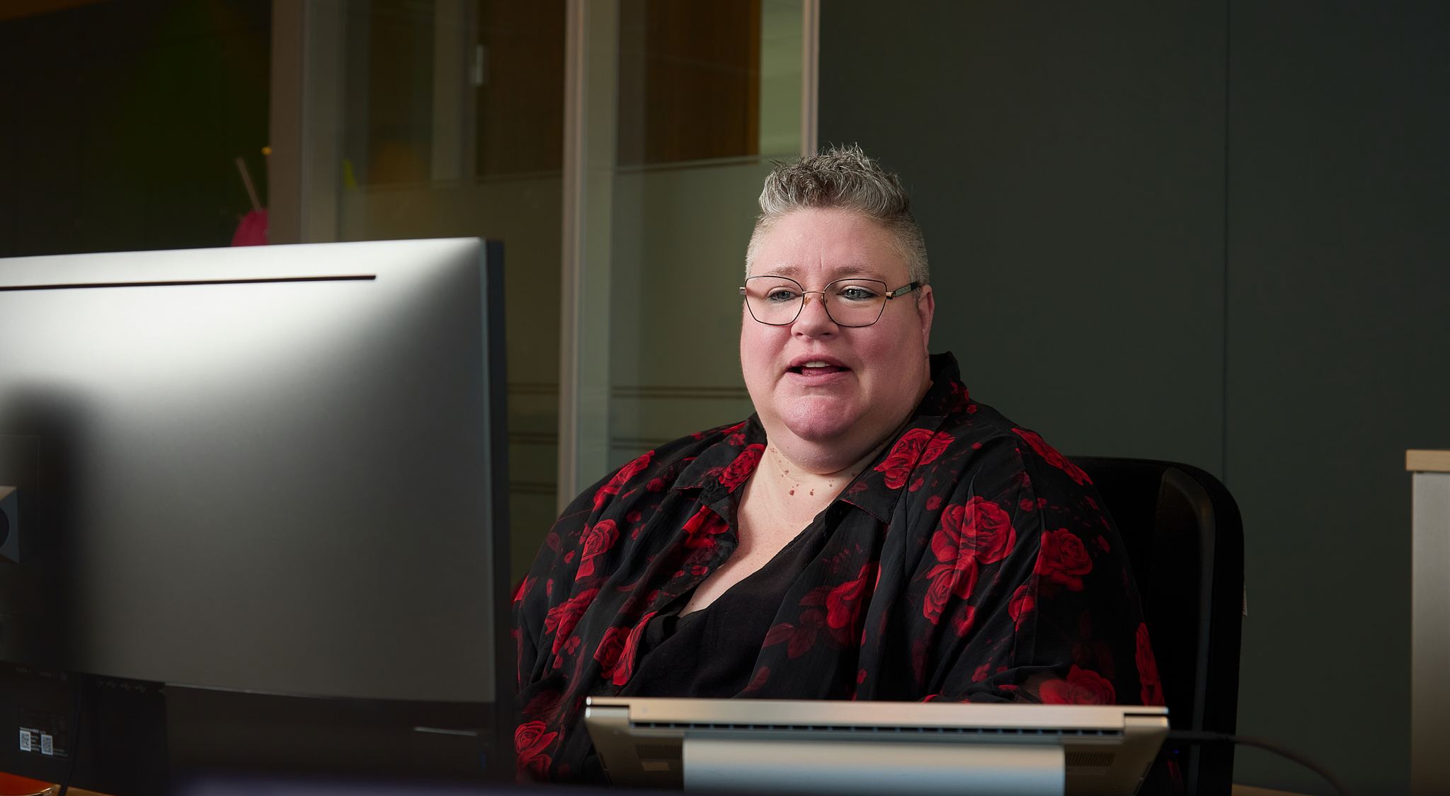 Person sitting at a desk working on a computer in an office setting, wearing glasses and a black shirt with a red floral pattern.