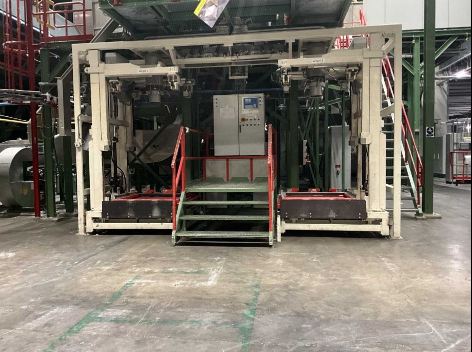 Industrial machine setup inside a factory, featuring metal framing with steps and railings leading to a control panel, surrounded by structural beams and other production equipment