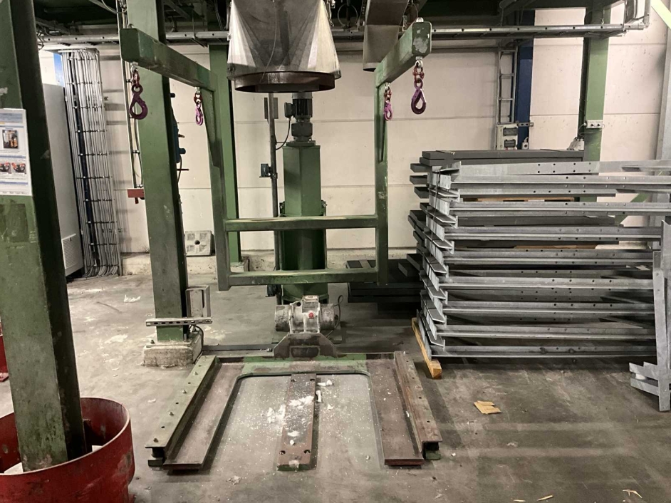Industrial equipment setup with a metal frame structure and funnel-shaped component, located in a workshop area with stacked metal parts on the right and exposed wiring on the left.
