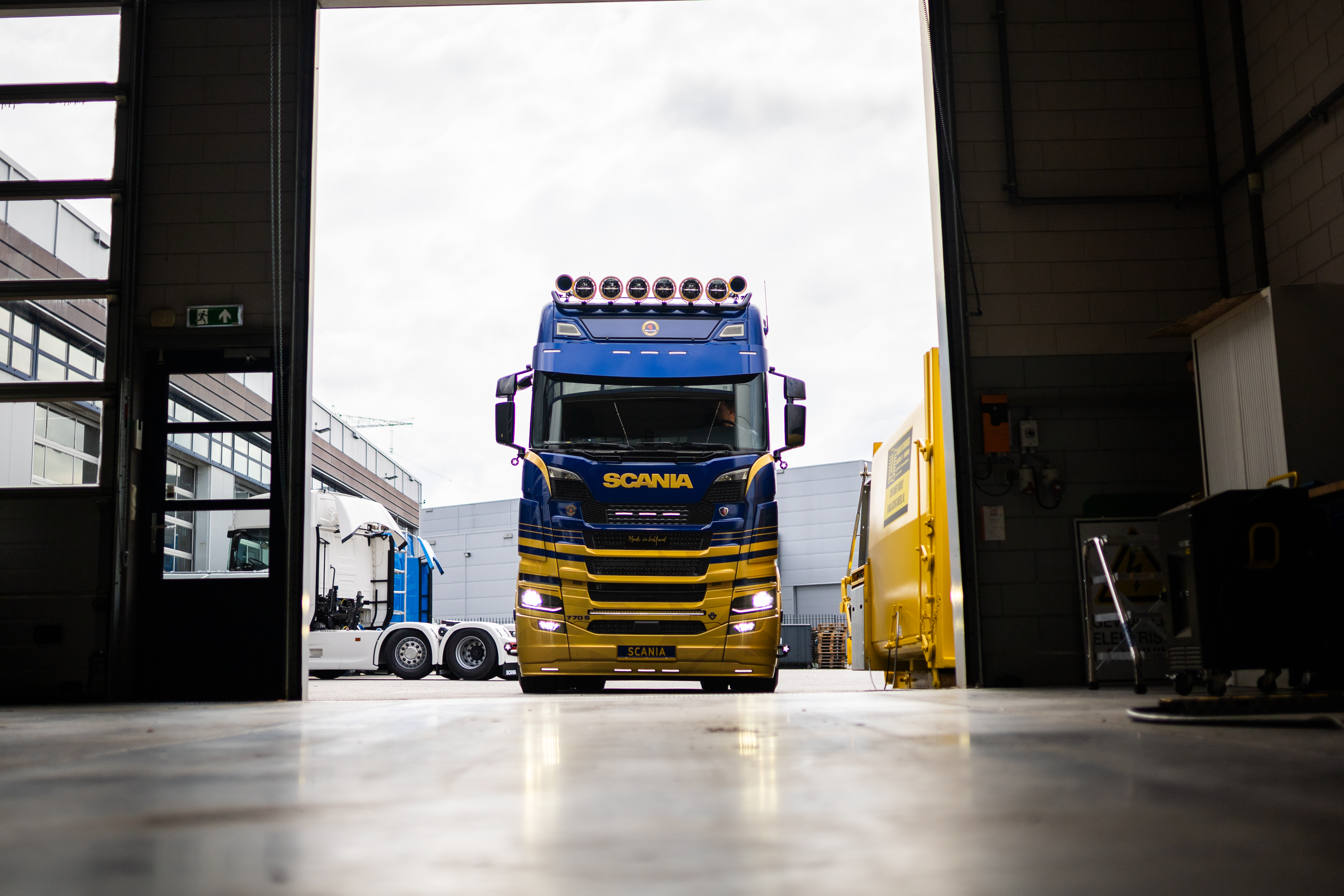 Blue and yellow Scania truck parked outside an industrial garage, viewed from inside the building with part of the workshop floor and doorway framing the scene.
