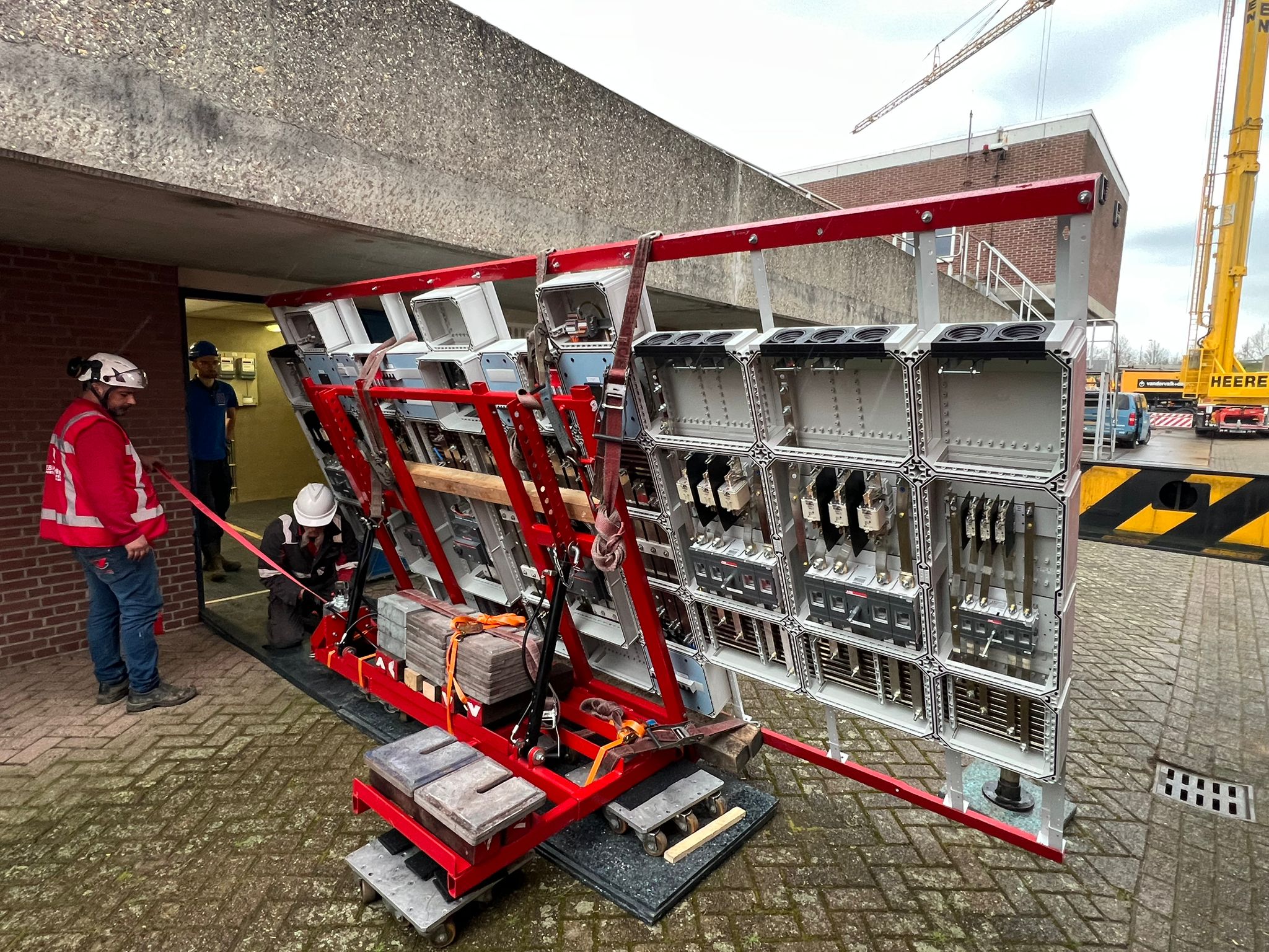 Werknemers in veiligheidskleding en helmen verplaatsen een groot industrieel elektrisch paneel op een rood hefraam buiten een betonnen gebouw, met een kraan en extra werknemers op de achtergrond.
