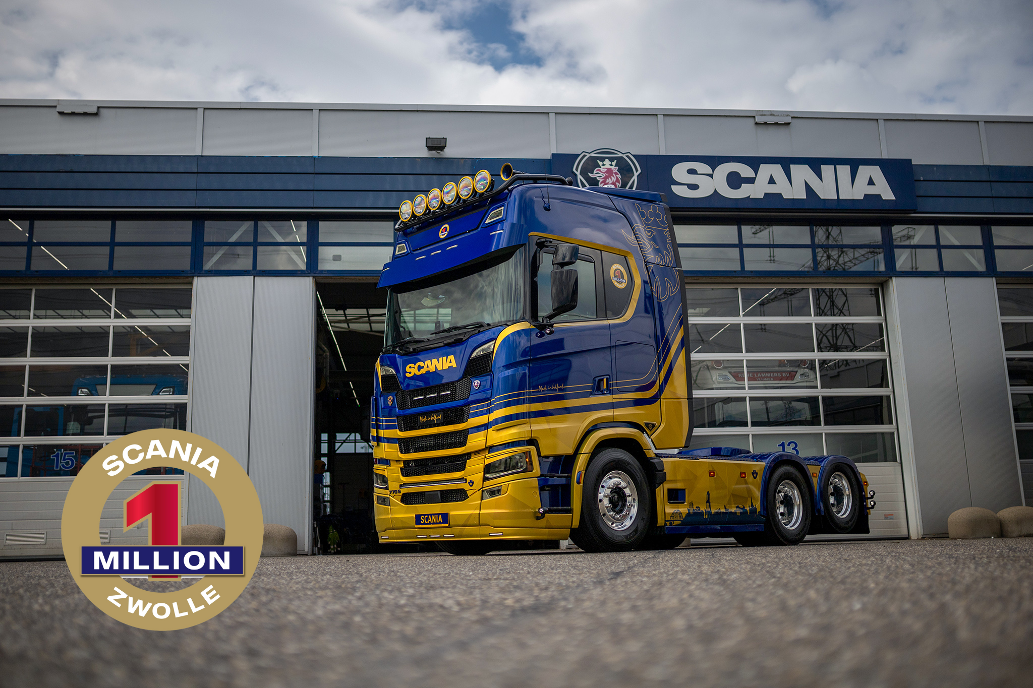 Blue and yellow Scania truck parked in front of a Scania service center building with large garage doors and the company logo above.