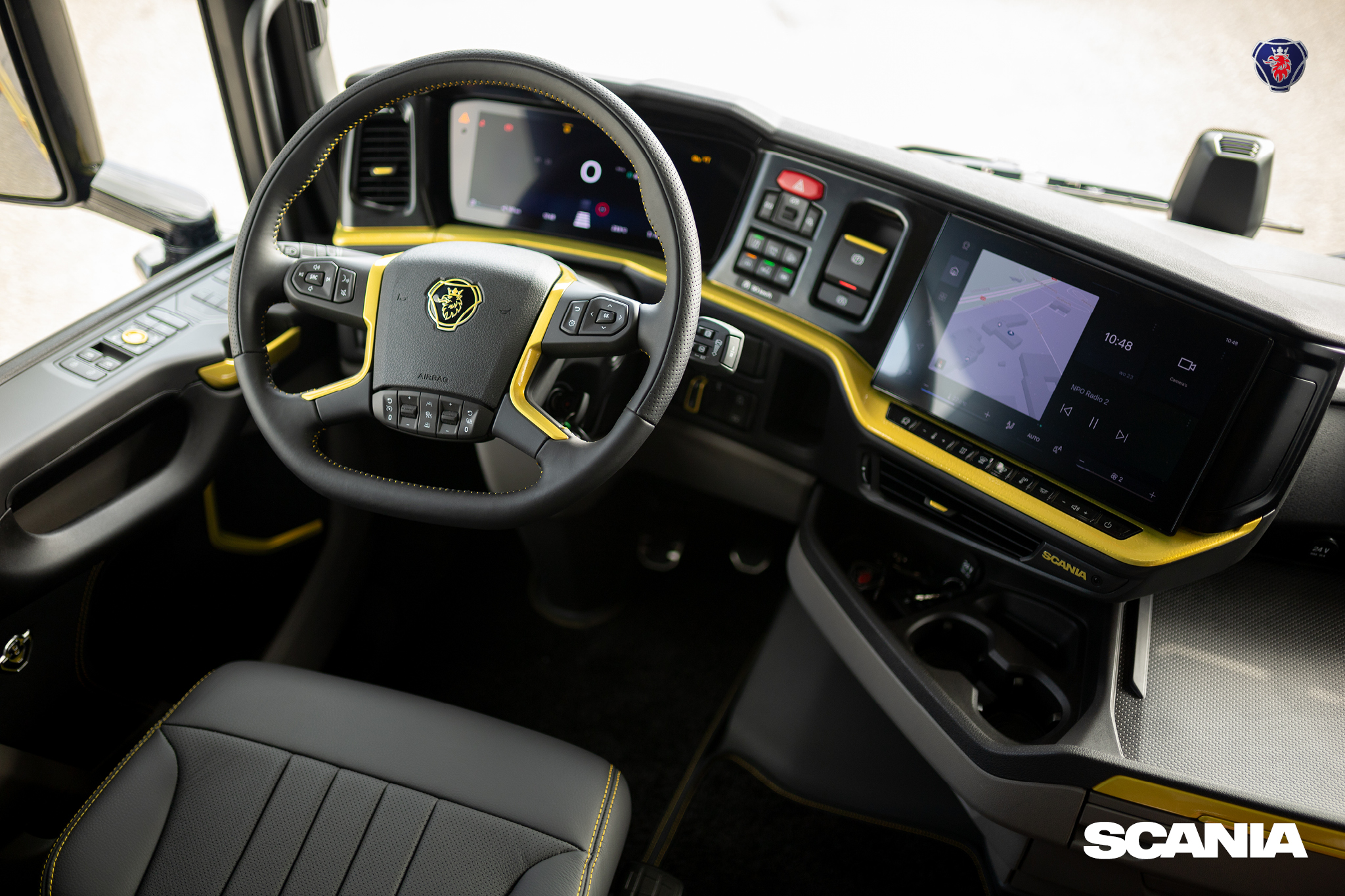 Interior view of a Scania truck cabin featuring a modern dashboard with touchscreen display, steering wheel with control buttons, and black and yellow detailing.