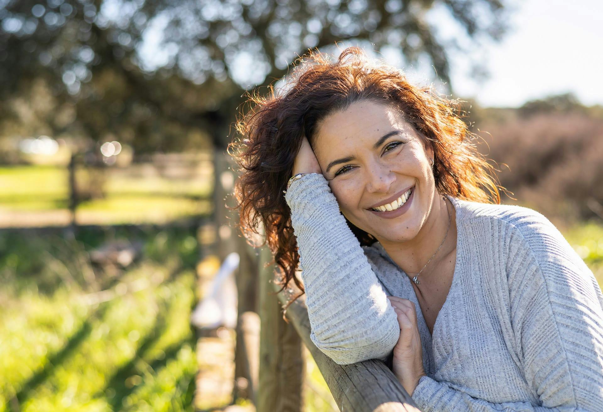 Smiling woman in grey sweater leaning on wooden fence.