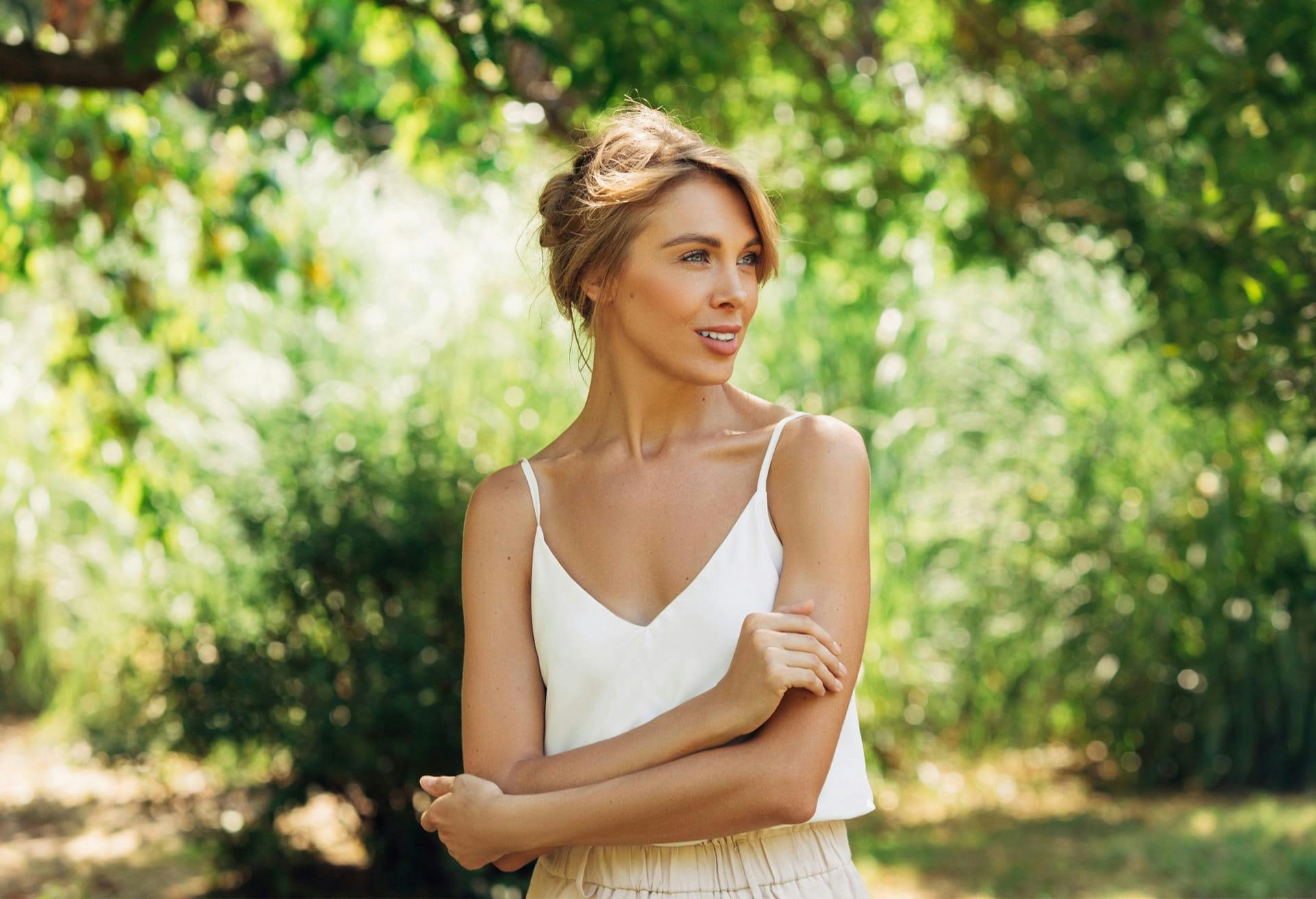 Woman in white blouse holding arms and looking to her left in front of trees.