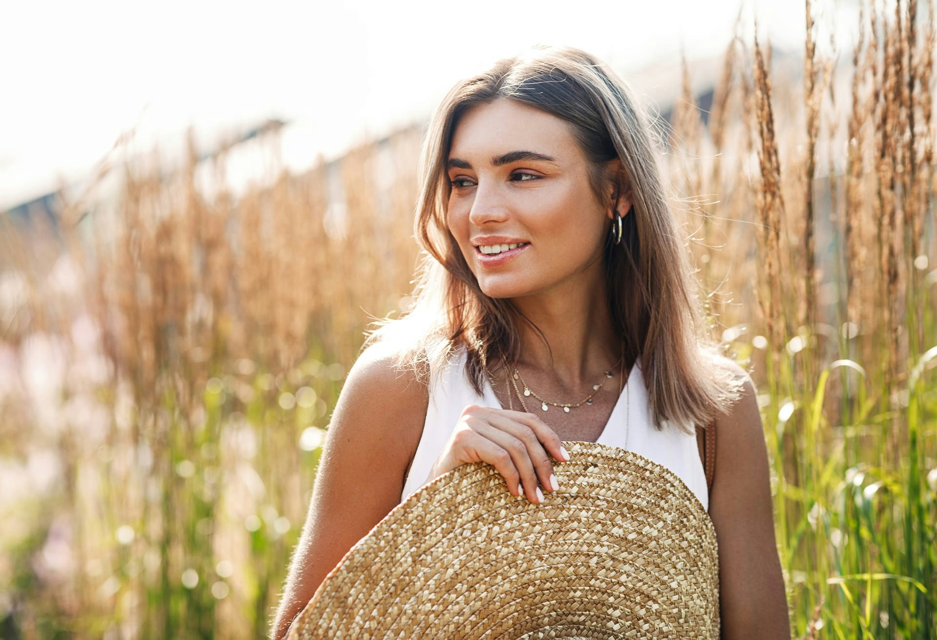 Smiling woman holding straw hat looking over right shoulder in front of wheat field.