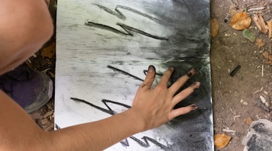 Ein Kind kniet auf dem Waldboden, vor ihm liegt ein Skizzenheft mit Zeichenpapier. Es zeichnet mit seinen Fingern und zerreibt das Kohelpulver auf dem Papier. Es zeichnet mit Kohlenstifte