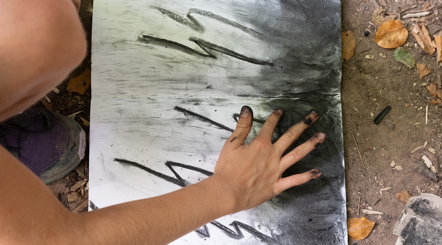 Ein Kind kniet auf dem Waldboden, vor ihm liegt ein Skizzenheft mit Zeichenpapier. Es zeichnet mit seinen Fingern und zerreibt das Kohelpulver auf dem Papier. Es zeichnet mit Kohlenstifte