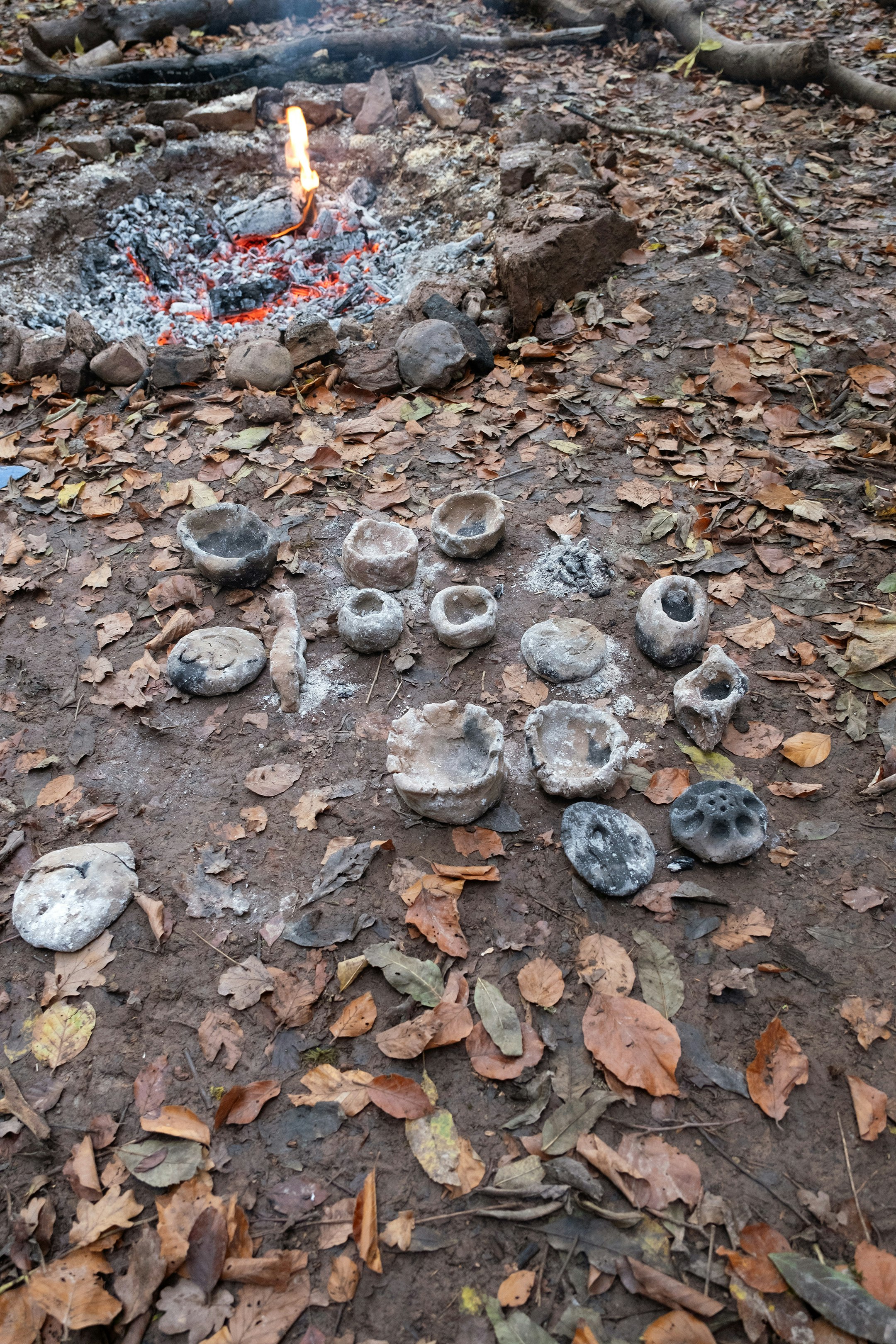 Ein Feuer brennt in einer Feuerstelle. Im Vordergrund sind mit Asche verschmierte Tongefässe auf den Waldboden gestellt.