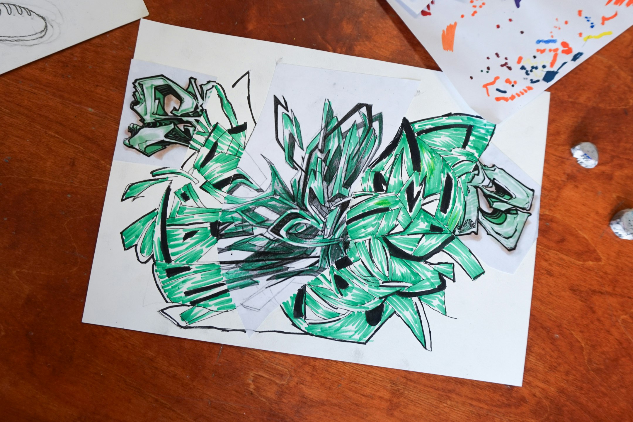 Ein Blatt Papier zeigt eine Skizze eines Graffitis. Es liegt auf einer hölzernen Untergrund. Die Skizze ist grün und schwarz.