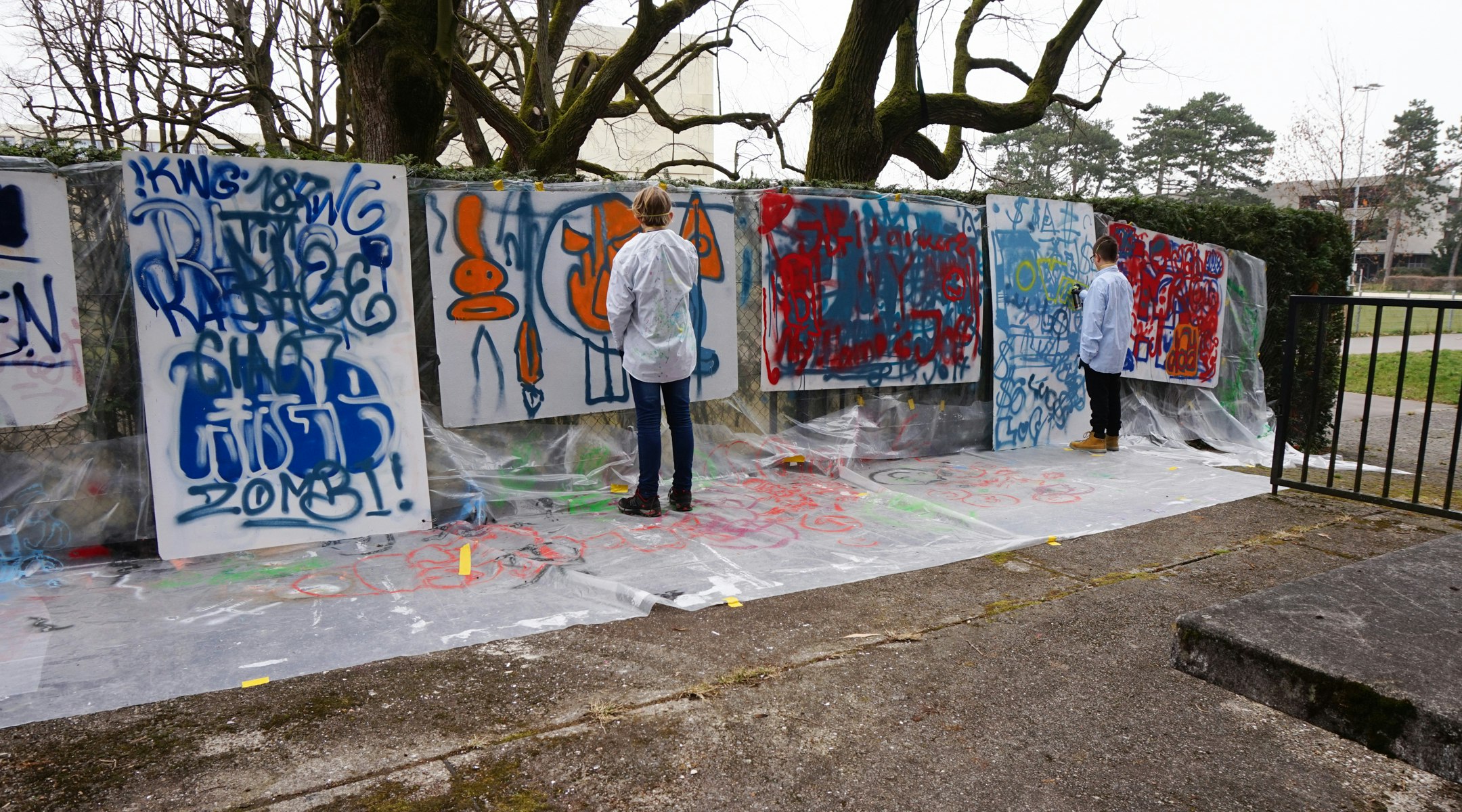 An einem Zaun hängen viele verschieden Papiere. Sie sind grossformatig. Dahinter ist ein Abdeckplastik. Auf den Papieren sind verschiedene Zeichnungen zu sehen, die mit der Spraydose gemacht wurden. Vor den Papieren stehen Kinder mit einer Maske vor dem Gesicht und einer Spraydose in der Hand.
