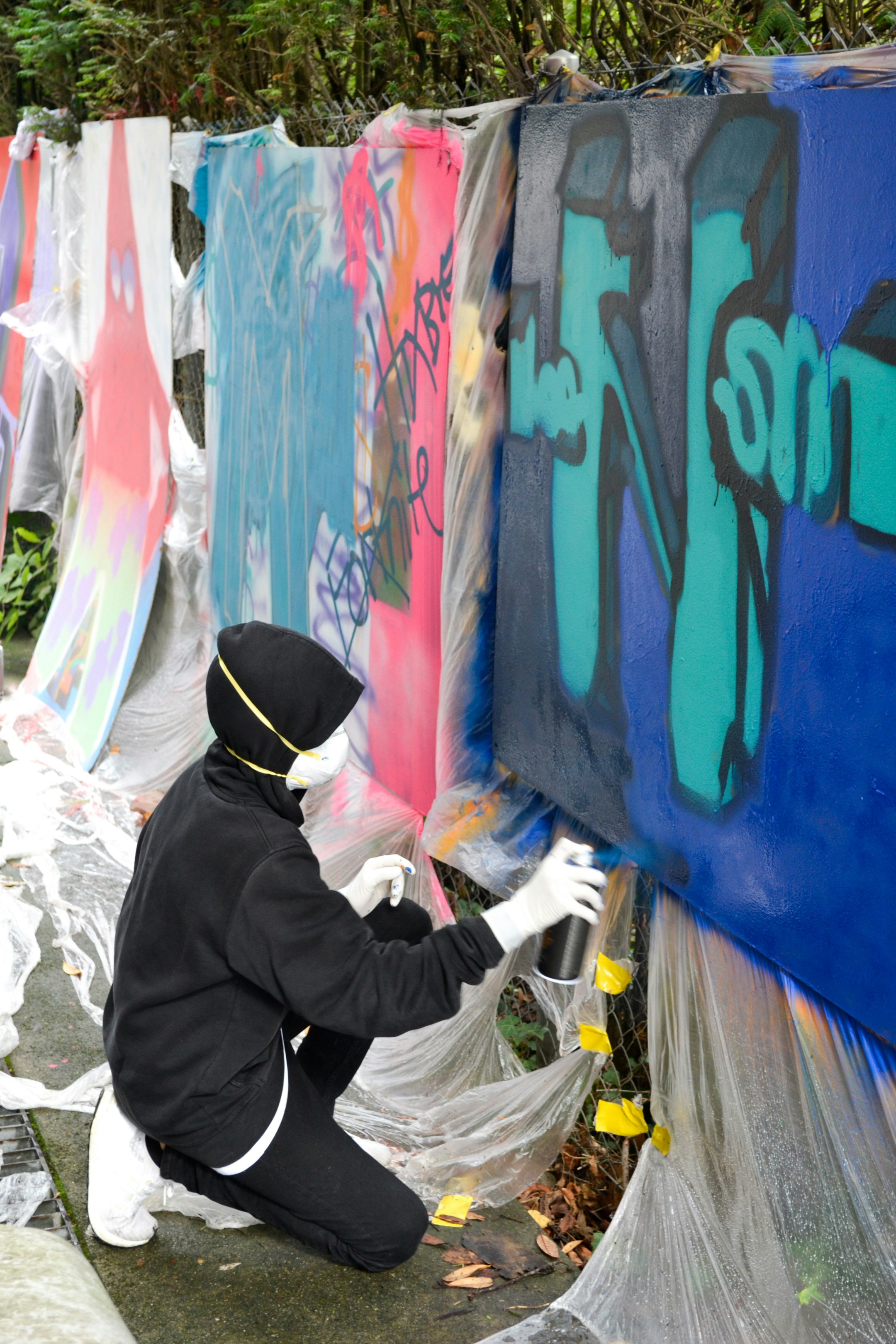 Eine jugendliche Person trägt eine Maske auf dem Gesicht und hält eine Spraydose in der Hand. Sie sprayt auf ein grossformatiges Papier, das vor ihr vertikal an einem Zaun befestigt ist.