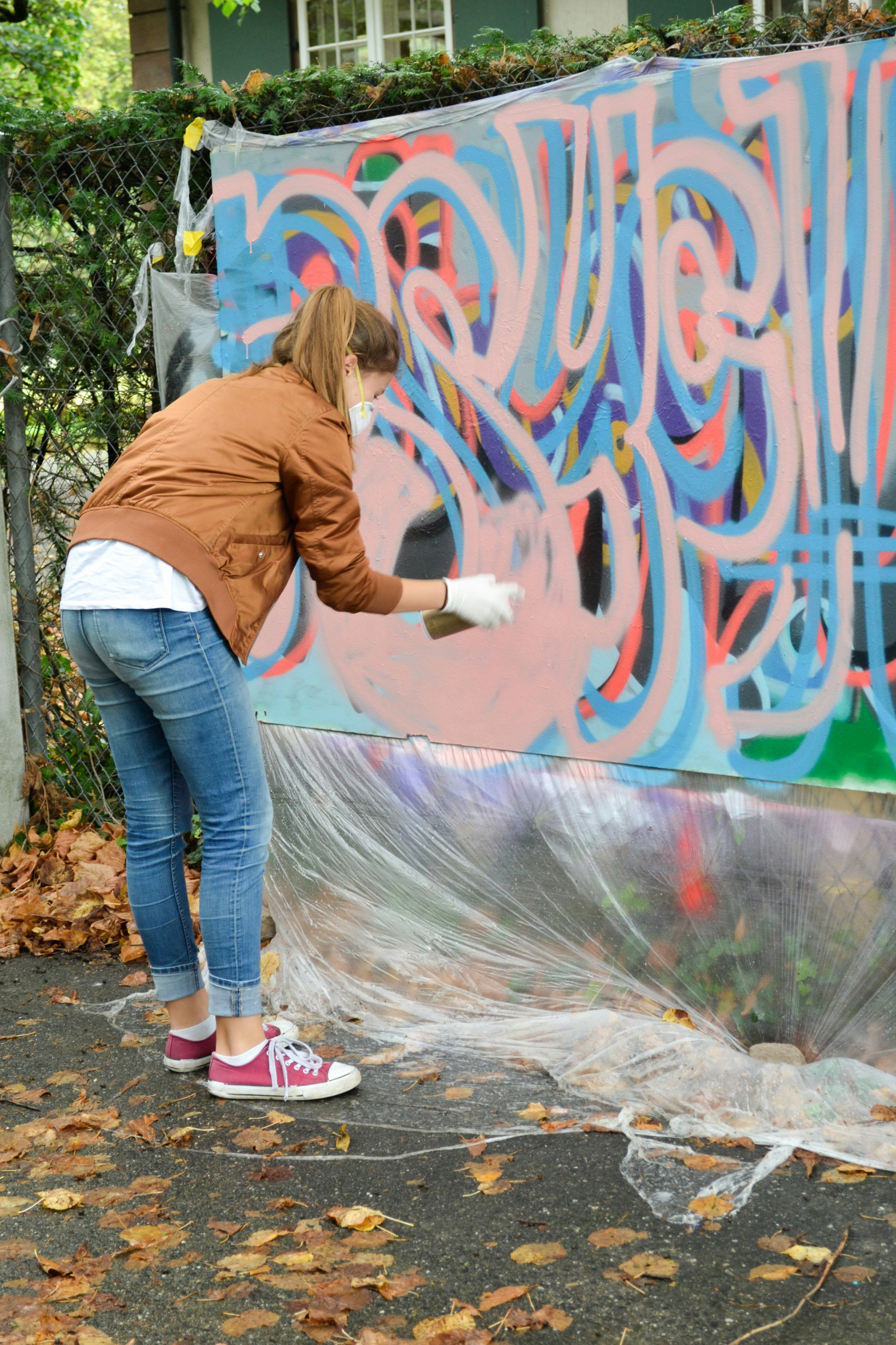 Ein Mädchen trägt eine Maske auf dem Gesicht und hält eine Spraydose in der Hand. Sie sprayt auf ein grossformatiges Papier, das vor ihr vertikal an einem Zaun befestigt ist.