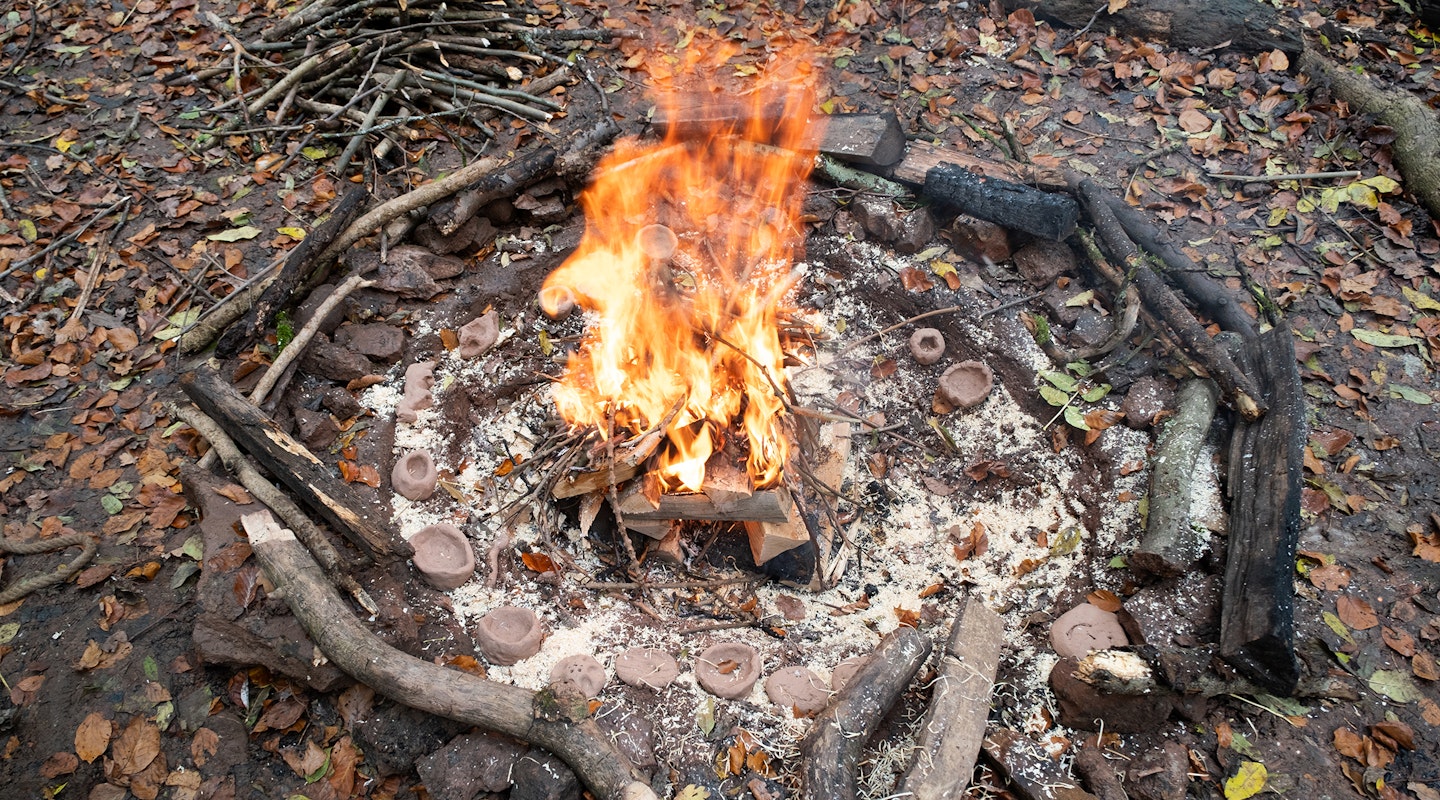 Ein Feuer im Wald. Es brennt und grosse Feuer sind sichtbar. In der Asche, rund um das Feuer verteilt, liegen selbst geformte Schalen aus rotem Ton.