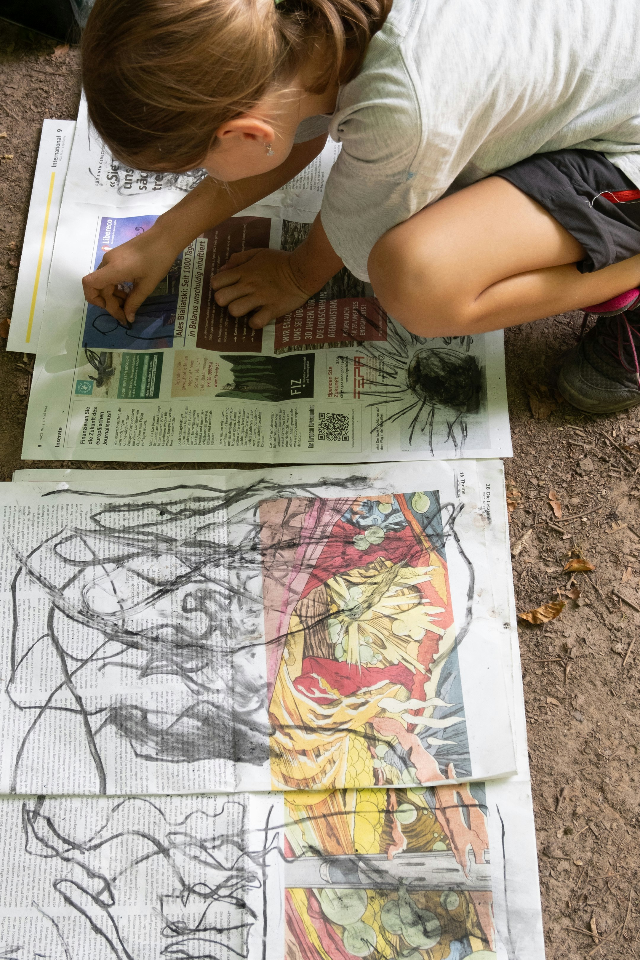 Ein Kind zeichnet mit einem Kohlenstift auf Zeitungspapier. Die Zeitung liegt aufgefaltet auf dem Waldboden. Das Kind kniet auf dem Waldboden.