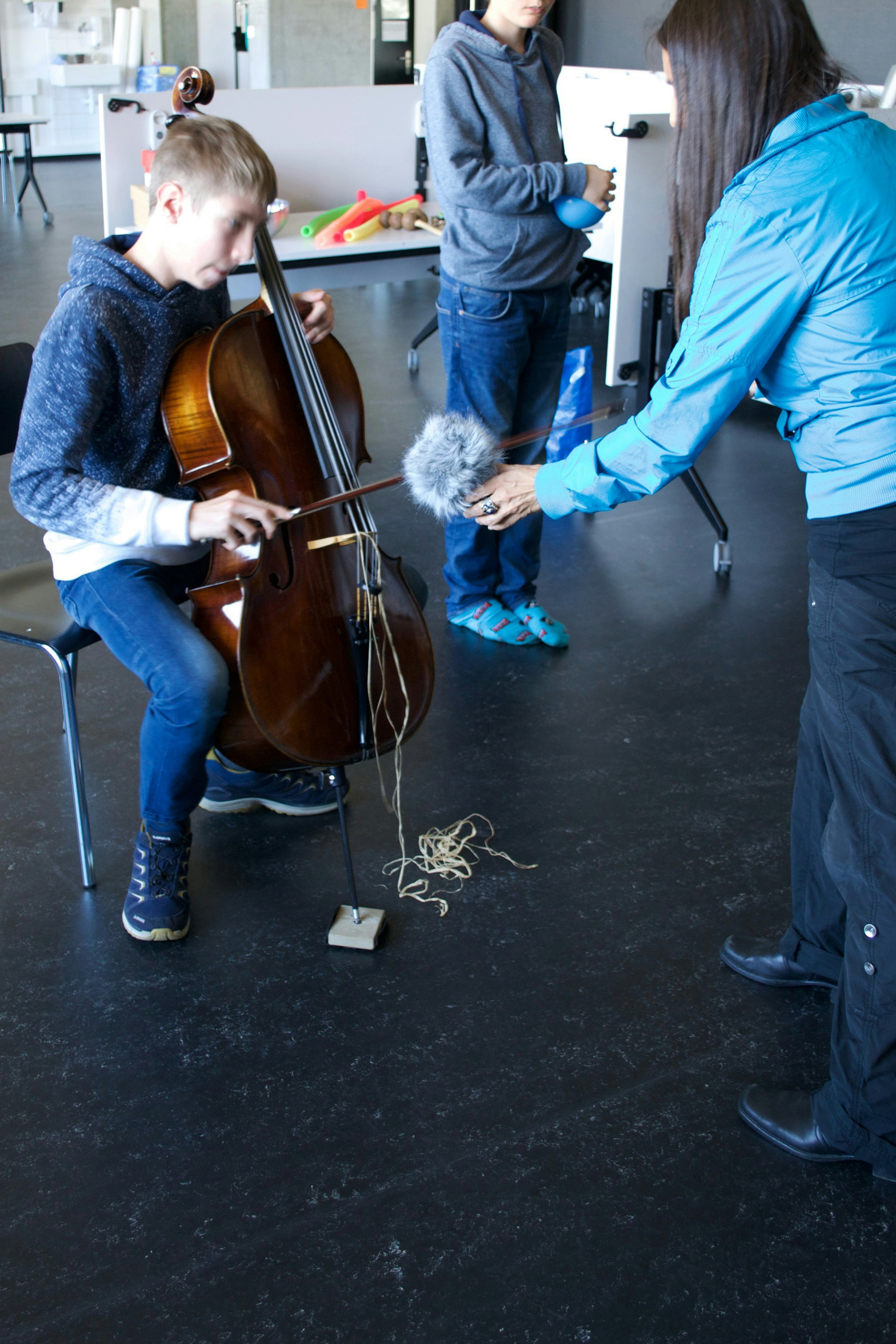 Eine Frau hält ein Audio-Aufnahmegerät in den Händen und hält es vor ein Cello. Ein Junge sitzt auf dem Stuhl und spielt Cello.
