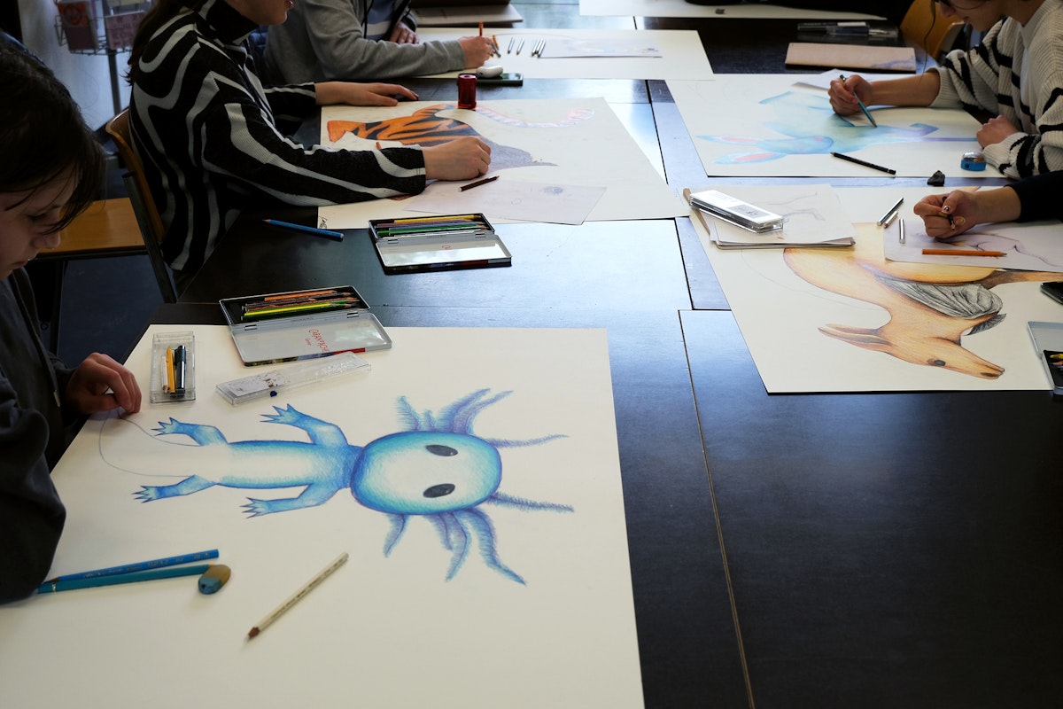 Kinder sitzen an einem Tisch und zeichnen mit Buntstiften auf A2-Papiere sehr unterschiedliche Fabelwesen, die sich aus Tier und märchenhafte Figuren zusammensetzen.
