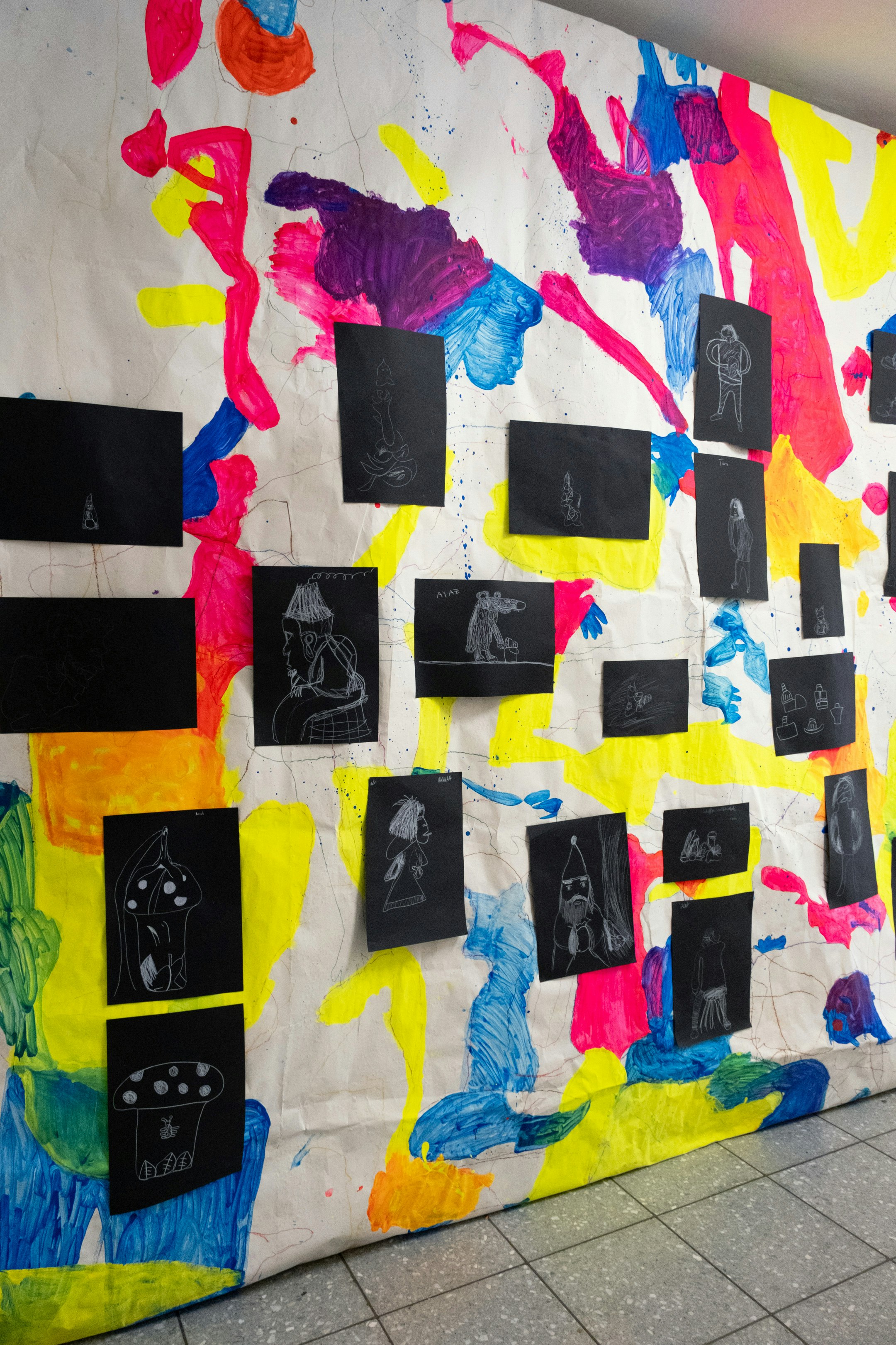 Ein grossformatiges Papier hängt an der Decke und reicht bis zum Boden. Verschiedene farbige und abstrakte Formen sind darauf gemalt. Schwarzweiss Zeichnungen von Kindern sind auf dem grossformatigem Papier angebracht.