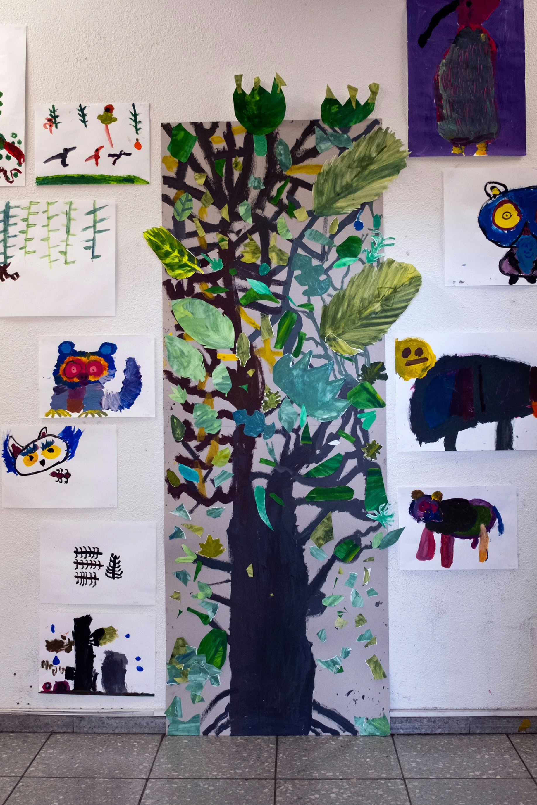 Malereien von Kindern auf Papier sind an einer Wand im Innenraum ausgestellt. In der Mitte ist ein Baum mit unterschiedlich grossen, grünen Blättern. Auf der Seite befinden sich verschiedenen Tiere.