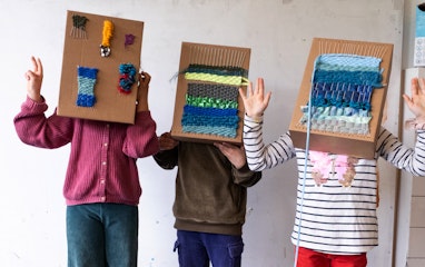Drei Kinder stehen mit Kartonschachteln über dem Kopf in einem Raum und gestikulieren mit den Händen. Die Kartonschachteln dienen als Webrahmen. Auf ihnen ist ein Webstück befestigt.