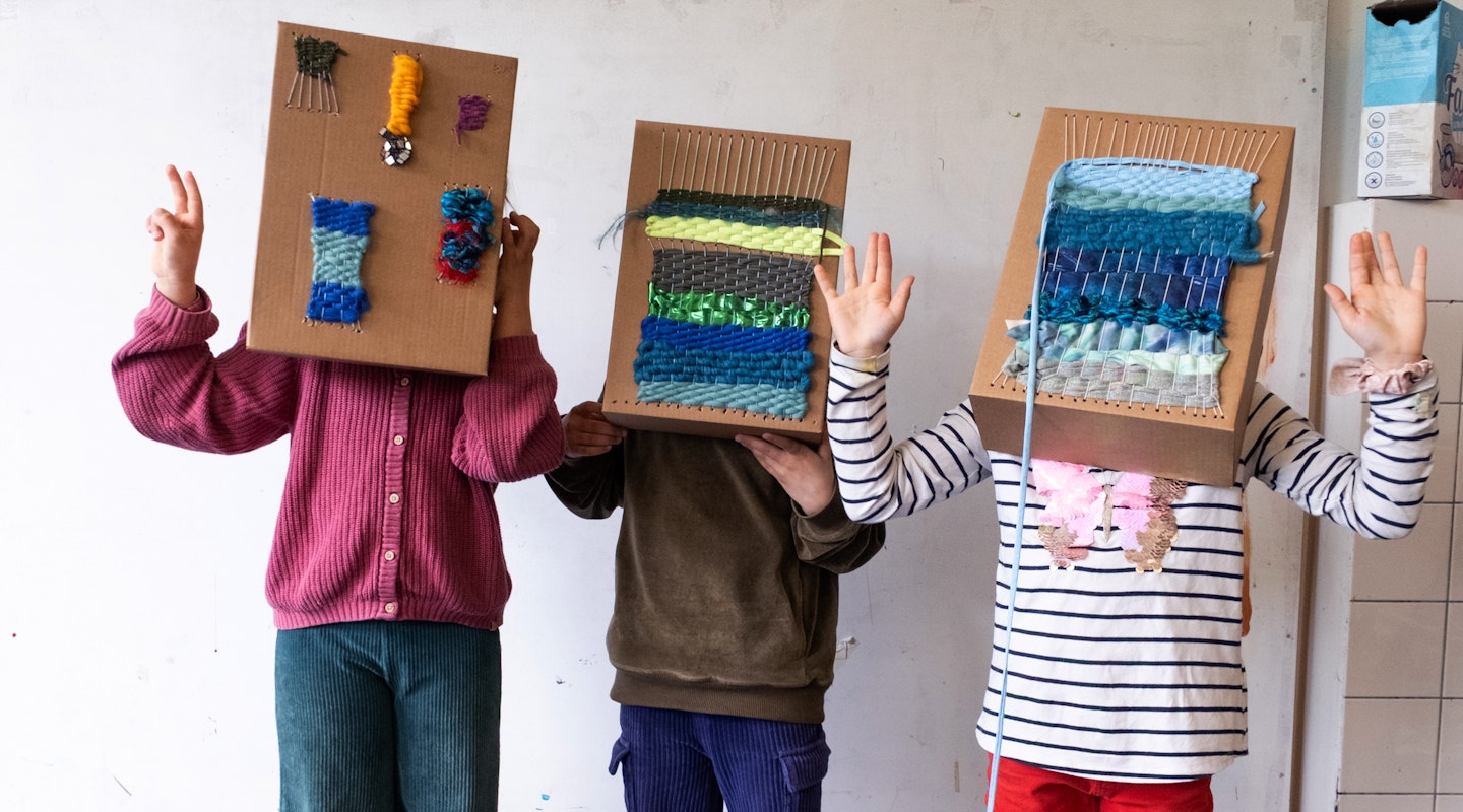 Drei Kinder stehen mit Kartonschachteln über dem Kopf in einem Raum und gestikulieren mit den Händen. Die Kartonschachteln dienen als Webrahmen. Auf ihnen ist ein Webstück befestigt.