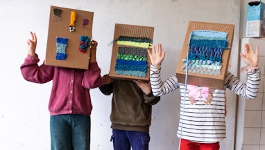 Drei Kinder stehen mit Kartonschachteln über dem Kopf in einem Raum und gestikulieren mit den Händen. Die Kartonschachteln dienen als Webrahmen. Auf ihnen ist ein Webstück befestigt.