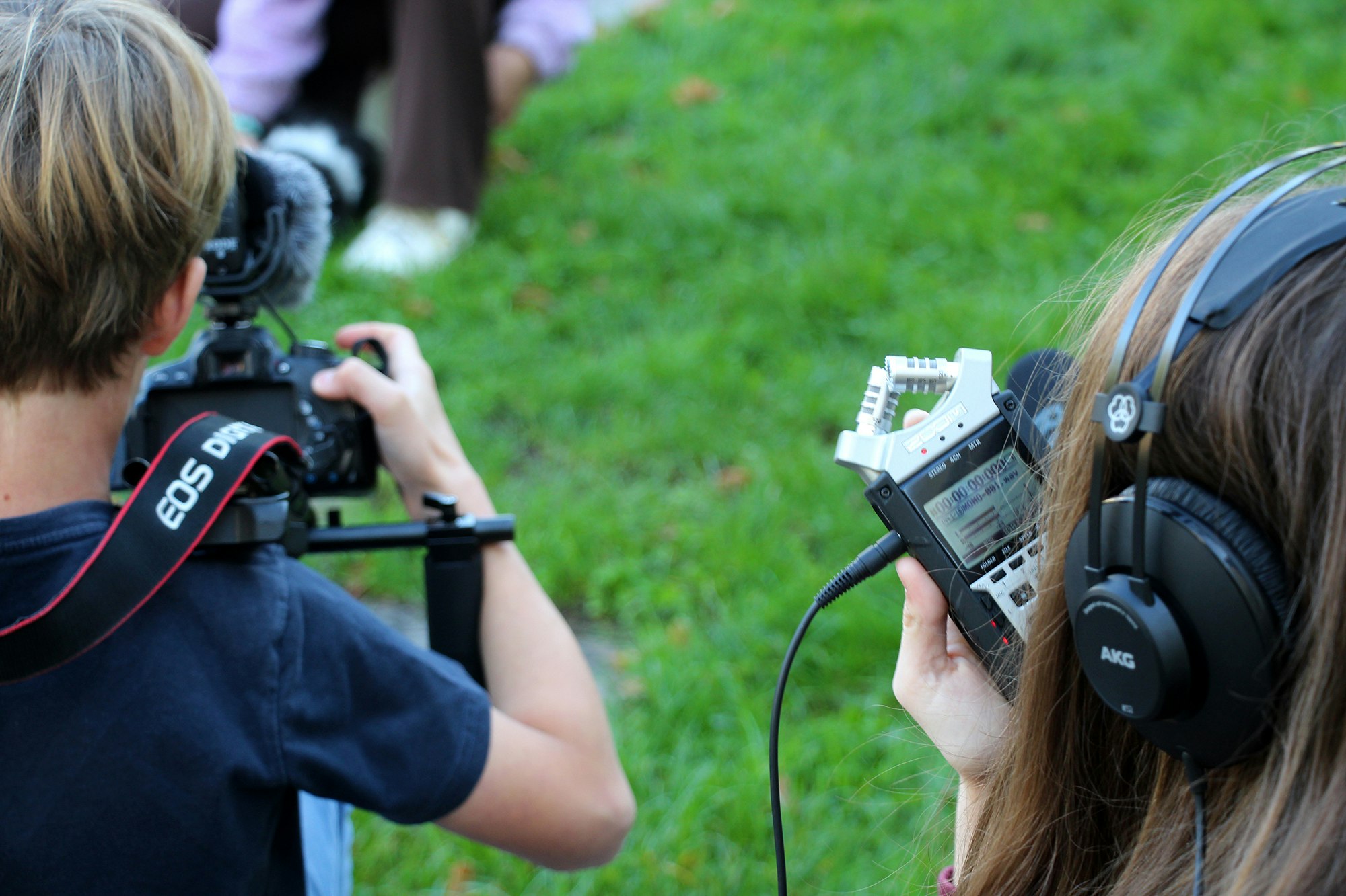 Zwei Kinder sind mit einer Kamera und einem Zoom-Aufnahmegerät ausgerüstet und sind draussen auf einer Wiese oder einem Stück Rasen unterwegs. Im Hintergrund – verschwommen sichtbar – bewegen sich andere Kinder.