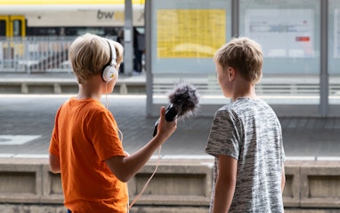Zwei Kinder stehen auf einem Perron neben dem Gleis. Ein Kind hält ein Audio-Aufnahmegerät in der Hand und einen Kopfhörer auf dem Kopf. Das andere Kind steht daneben und spricht etwas ins Audio-Aufnahmegerät.