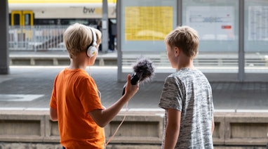 Zwei Kinder stehen auf einem Perron neben dem Gleis. Ein Kind hält ein Audio-Aufnahmegerät in der Hand und einen Kopfhörer auf dem Kopf. Das andere Kind steht daneben und spricht etwas ins Audio-Aufnahmegerät.