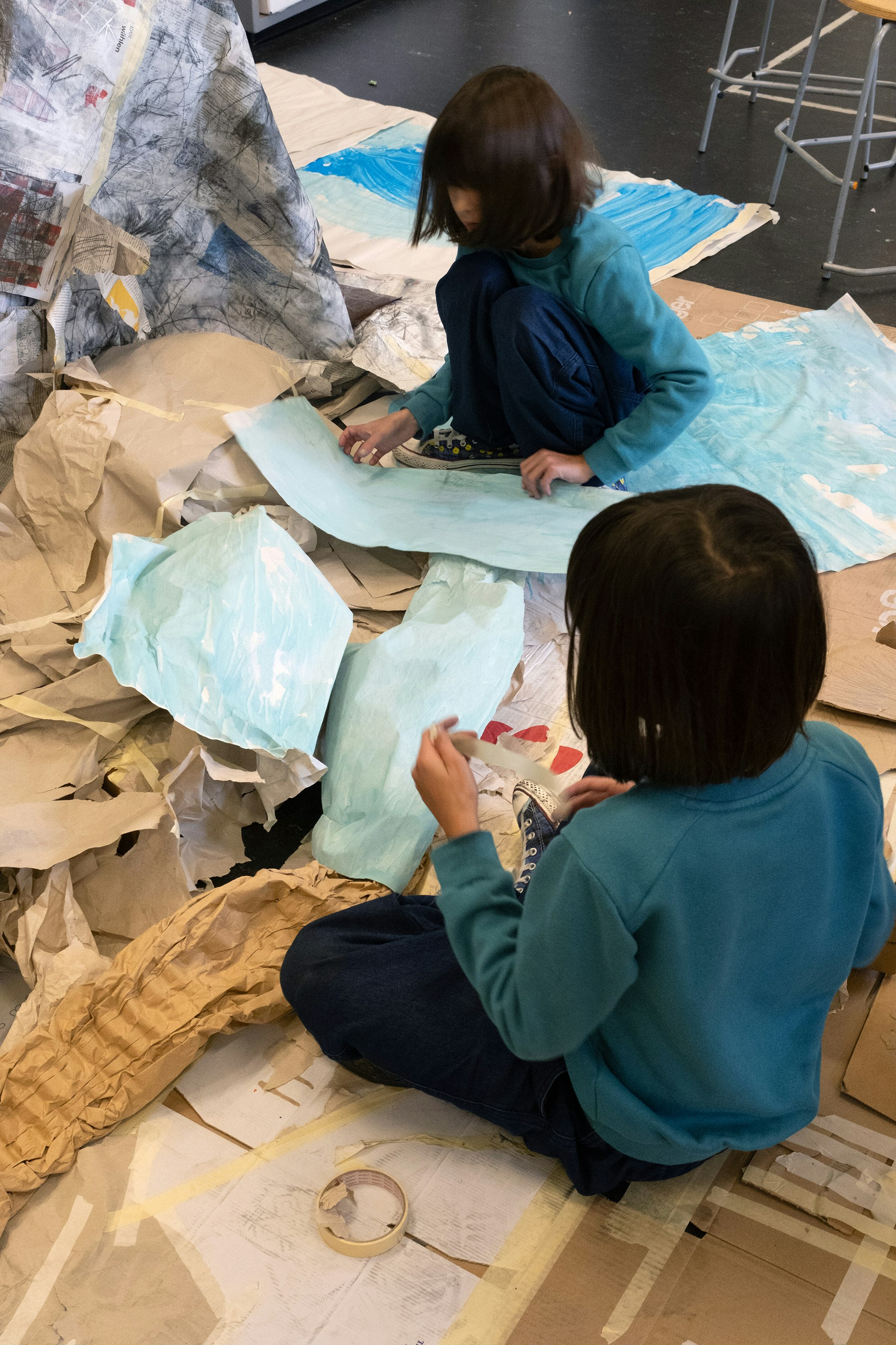 Zwei Kinder sitzen in mitten von grossen Papier- und Kartonstücken am Boden. Sie halten Klebestreifen in ihren Händen und Kleben einzelne Papierstücke aneinander.