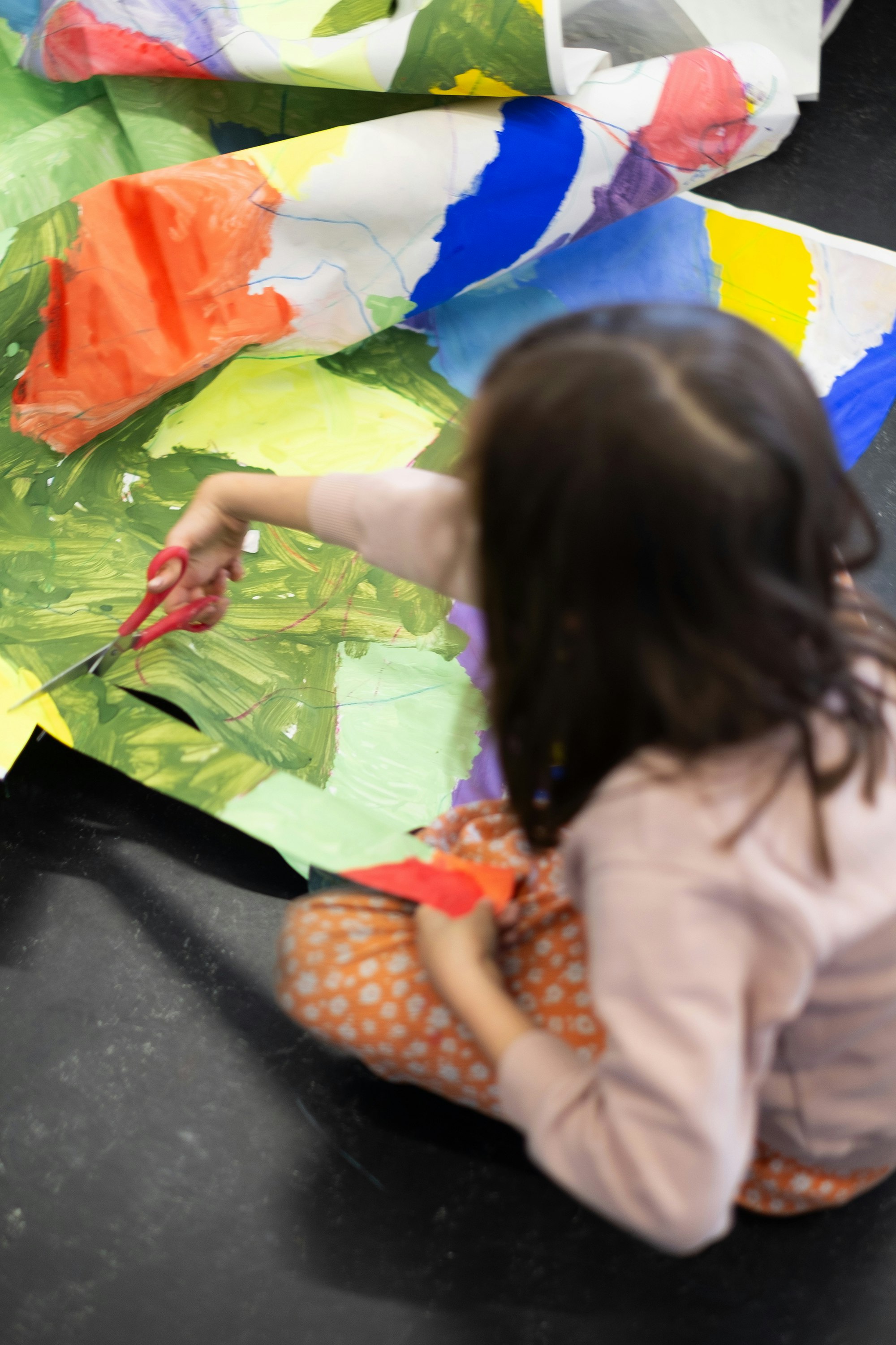 Ein Kind sitzt am Boden. Es hält eine Schere in der Hand und zerschneidet ein grosses, bemaltes Papier. EIne Szene aus dem K'Werk Workshop «Unser Welt aus Papier», geleitet von Celia Sidler und Sonja Lippuer.