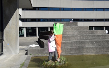 Ein Kind hält ein Papierobjekt. Das Papierobjekt stellt eine Karrotte dar, die so gross ist, wie das Kind selbst. Das Kind steht auf einem kleinen Inselchen inmitten des Teiches auf dem Pausenhof der Schule für Gestaltung Basel.