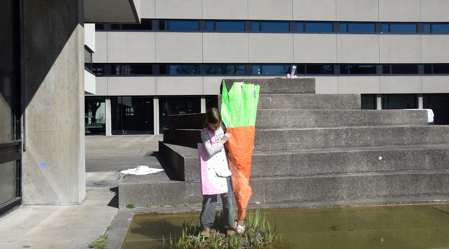 Ein Kind hält ein Papierobjekt. Das Papierobjekt stellt eine Karrotte dar, die so gross ist, wie das Kind selbst. Das Kind steht auf einem kleinen Inselchen inmitten des Teiches auf dem Pausenhof der Schule für Gestaltung Basel.