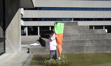 Ein Kind hält ein Papierobjekt. Das Papierobjekt stellt eine Karrotte dar, die so gross ist, wie das Kind selbst. Das Kind steht auf einem kleinen Inselchen inmitten des Teiches auf dem Pausenhof der Schule für Gestaltung Basel.