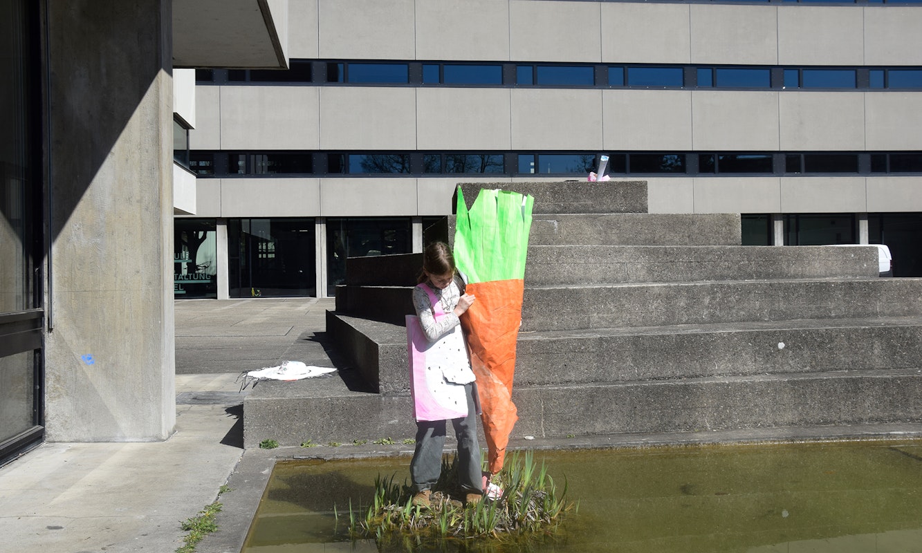 Ein Kind hält ein Papierobjekt. Das Papierobjekt stellt eine Karrotte dar, die so gross ist, wie das Kind selbst. Das Kind steht auf einem kleinen Inselchen inmitten des Teiches auf dem Pausenhof der Schule für Gestaltung Basel.