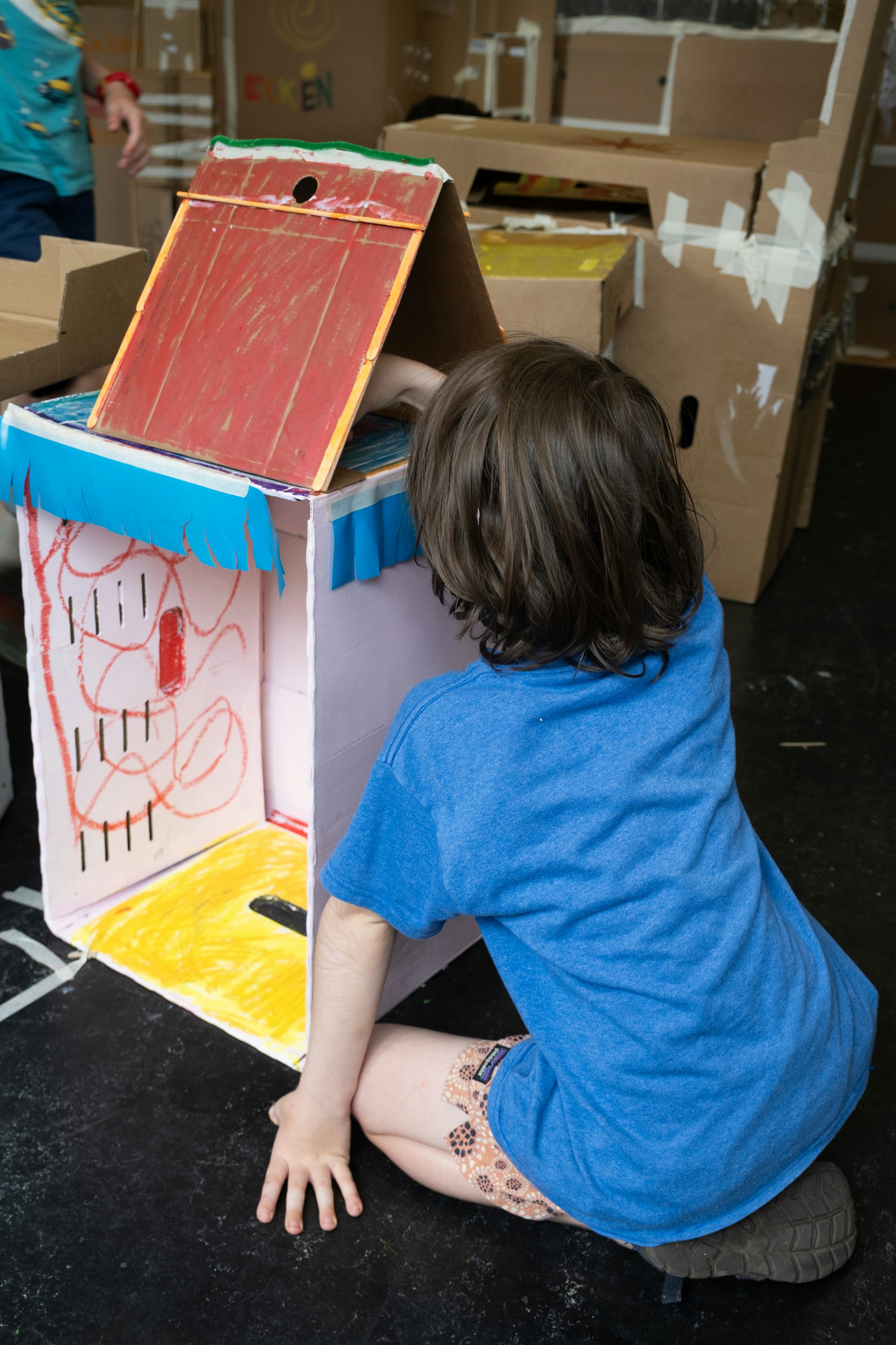 Ein Kind sitzt vor einem Kartonobjekt. Das Kartonobjekt wurde selbst gebaut. Es besteht aus Karton. Der Karton wurde bemalt.