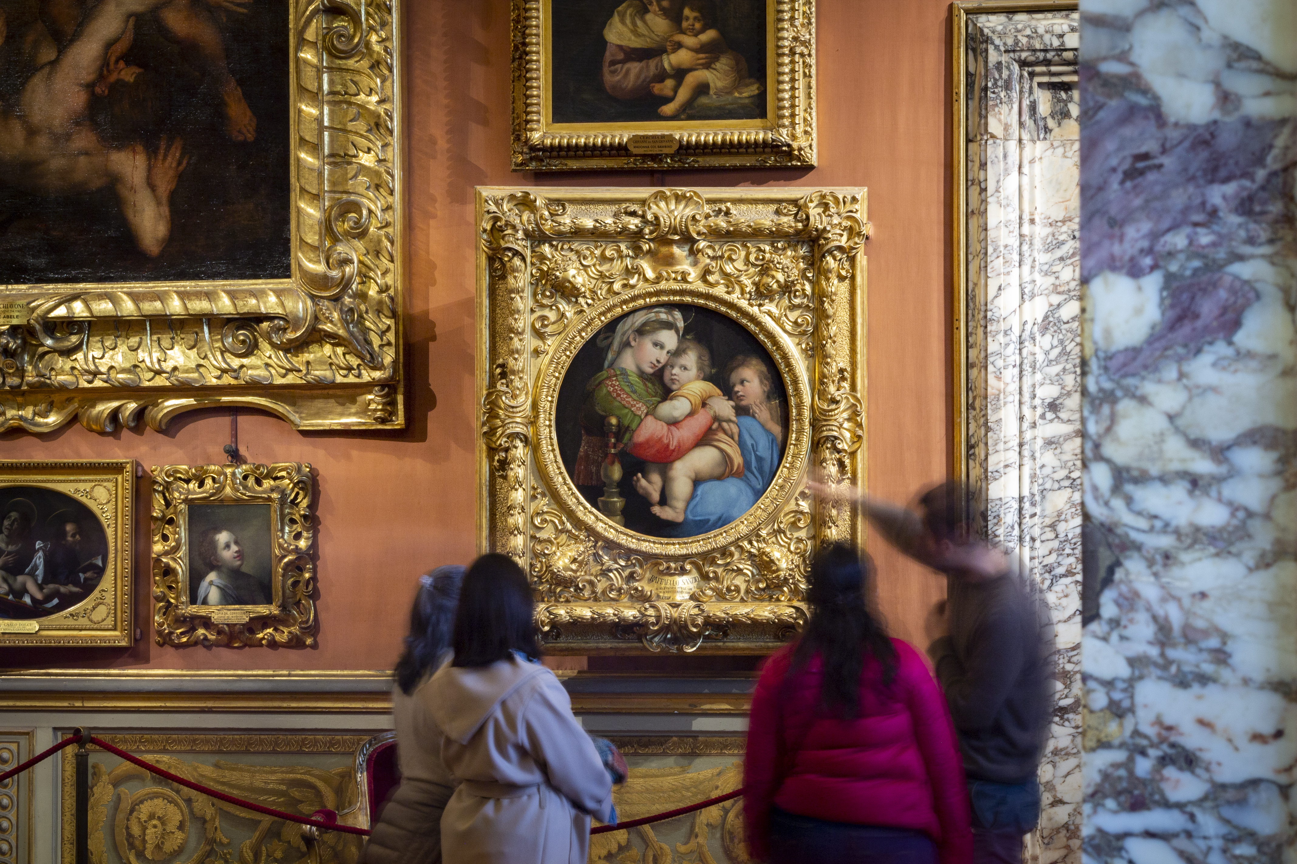 Un gruppo di persone osserva una collezione di quadri in una galleria d'arte elegantemente decorata, con un dipinto principale incorniciato in oro al centro della parete.