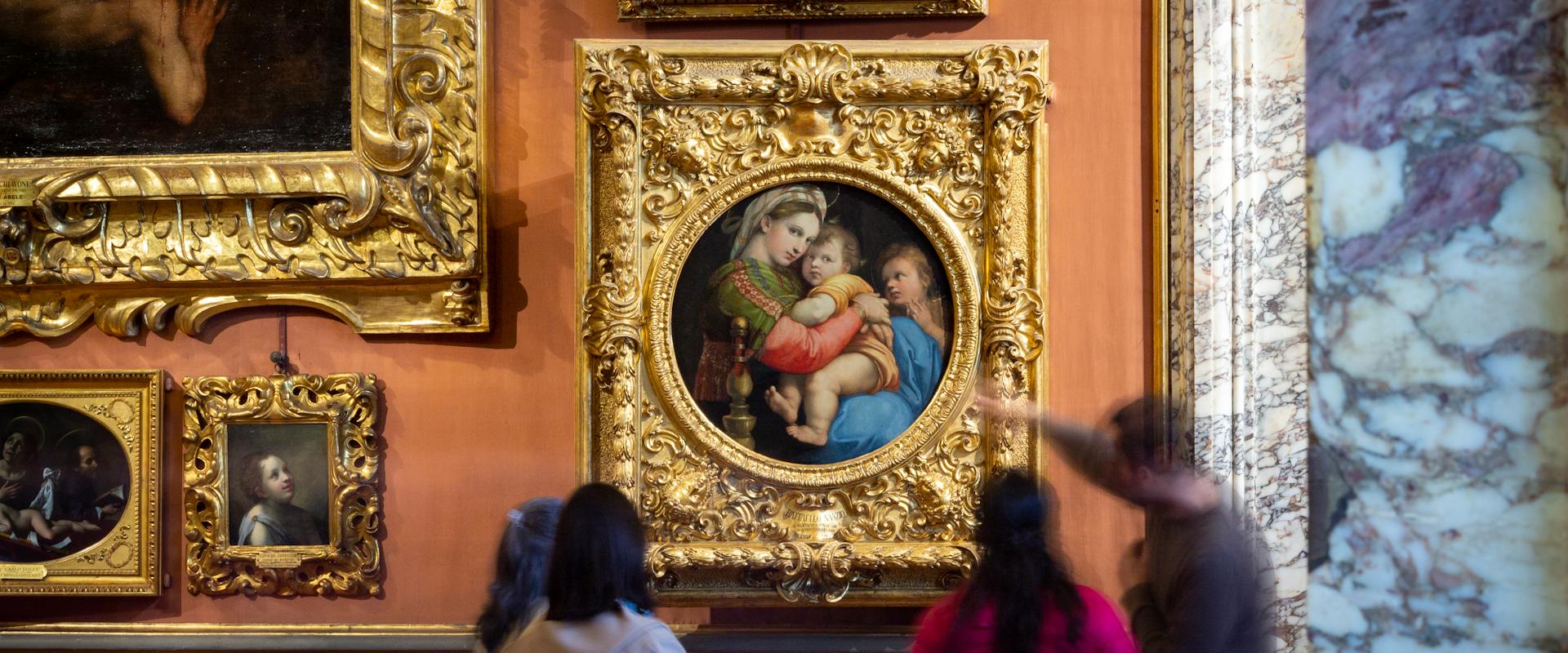 Un gruppo di persone osserva una collezione di quadri in una galleria d'arte elegantemente decorata, con un dipinto principale incorniciato in oro al centro della parete.