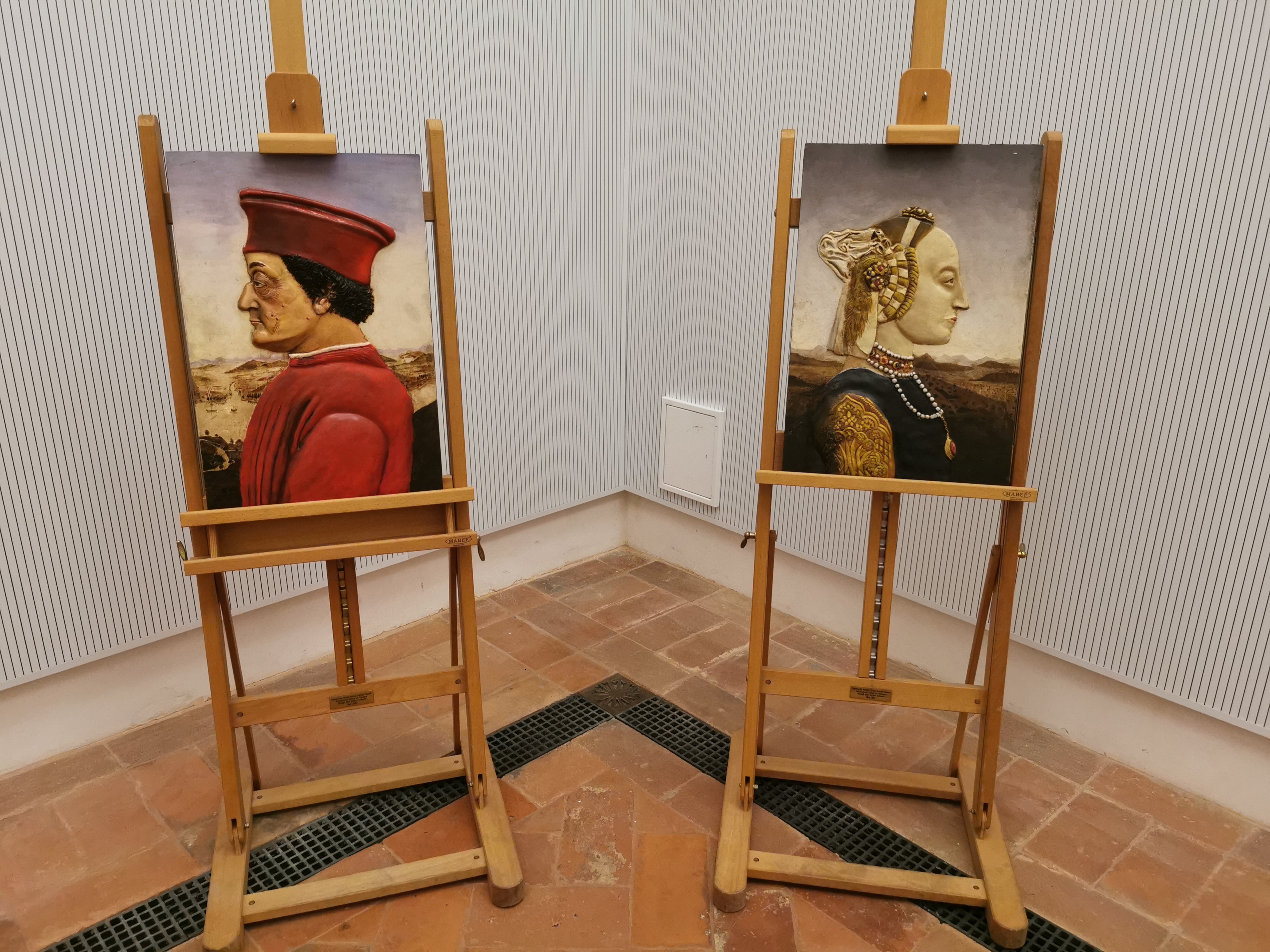 Due cavalletti in uno spazio espositivo mostrano ritratti rinascimentali di profilo, con un uomo in abiti rossi a sinistra e una donna dai elaborati ornamenti a destra, su pavimento in cotto.