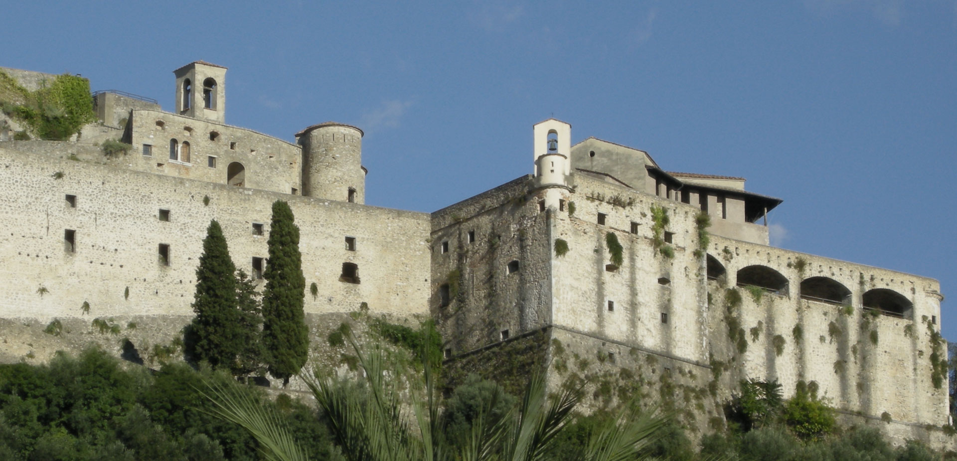 Il Castello di Itri si erge maestoso contro un cielo azzurro, con antiche mura in pietra e torri storiche circondate da vegetazione rigogliosa.