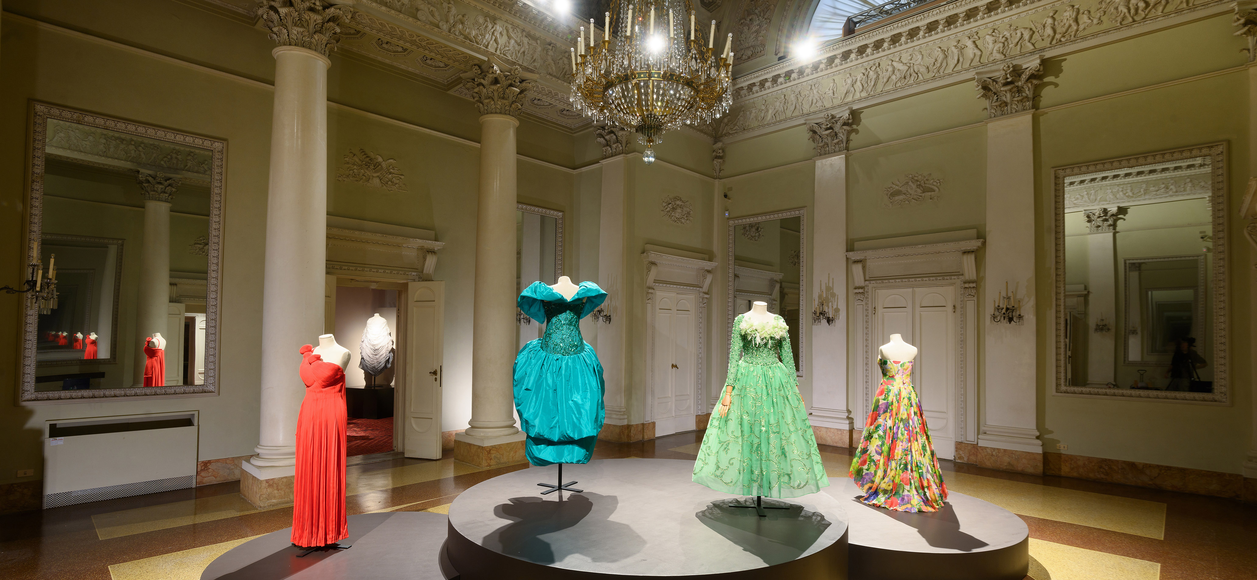 Nell'immagine si trova una sala espositiva lussuosa con alti soffitti ornati, in cui manichini indossano abiti eleganti e colorati in un ambiente raffinato con pavimento in marmo e grandi specchi.