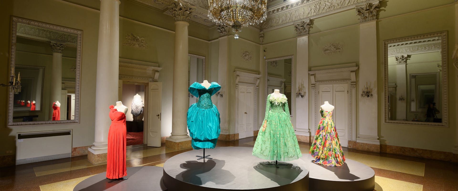 Nell'immagine si trova una sala espositiva lussuosa con alti soffitti ornati, in cui manichini indossano abiti eleganti e colorati in un ambiente raffinato con pavimento in marmo e grandi specchi.