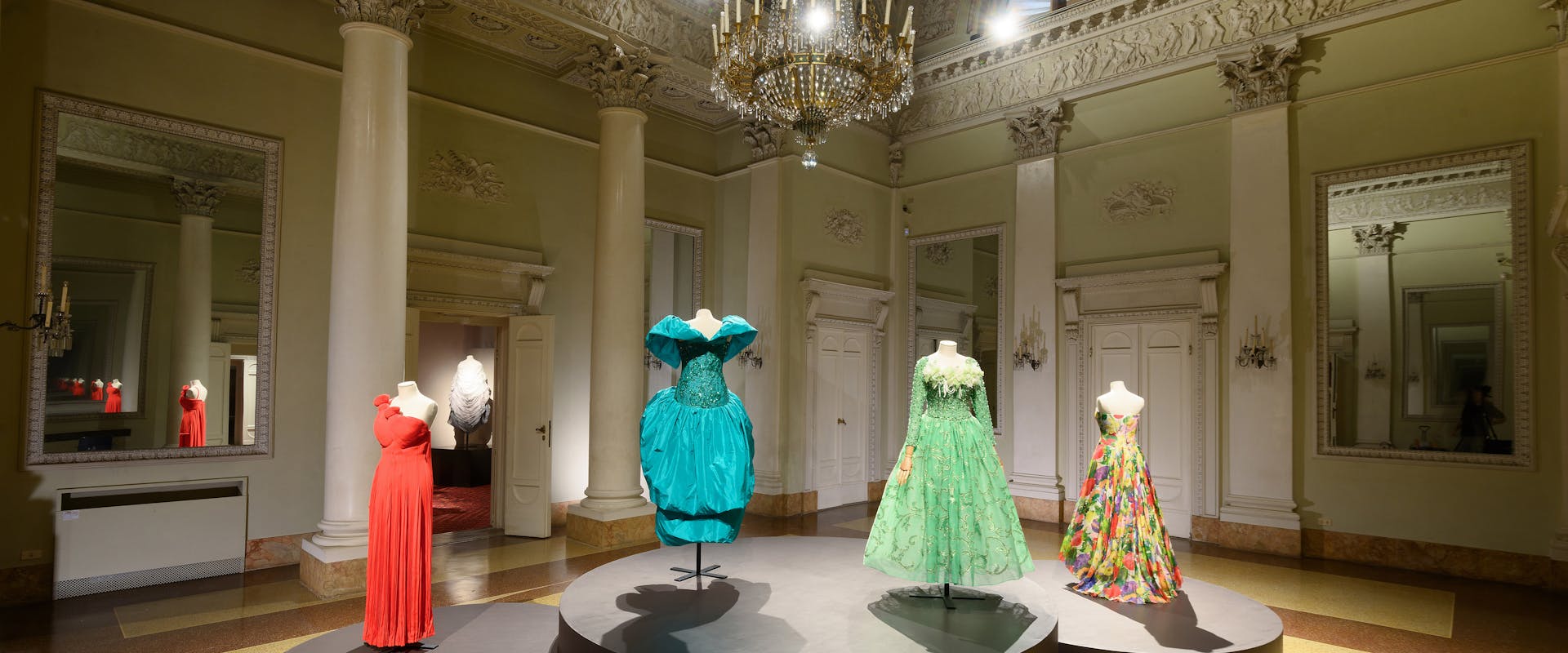 Luxurious ballroom displaying four elegant couture gowns on mannequins under a grand chandelier, featuring intricate designs in vibrant red, teal, green, and multi-colored patterns, set against a backdrop of ornate architectural details and large mirrors.