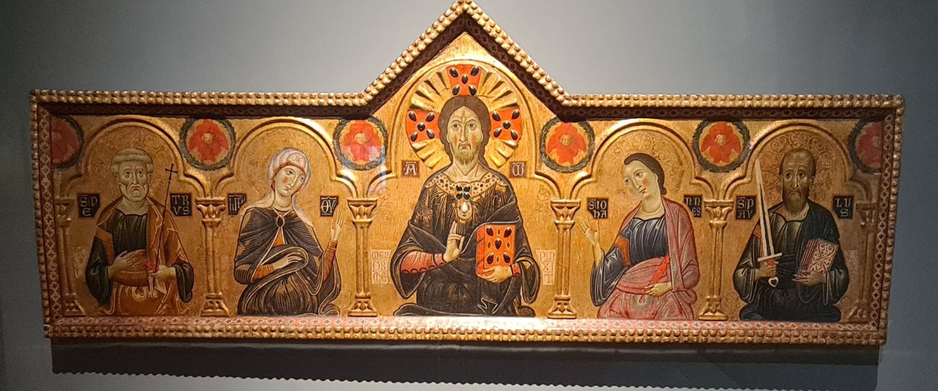 Un'antica pala d'altare medievale mostra figure sacre in abiti tradizionali, incorniciate da archi decorativi su uno sfondo dorato.