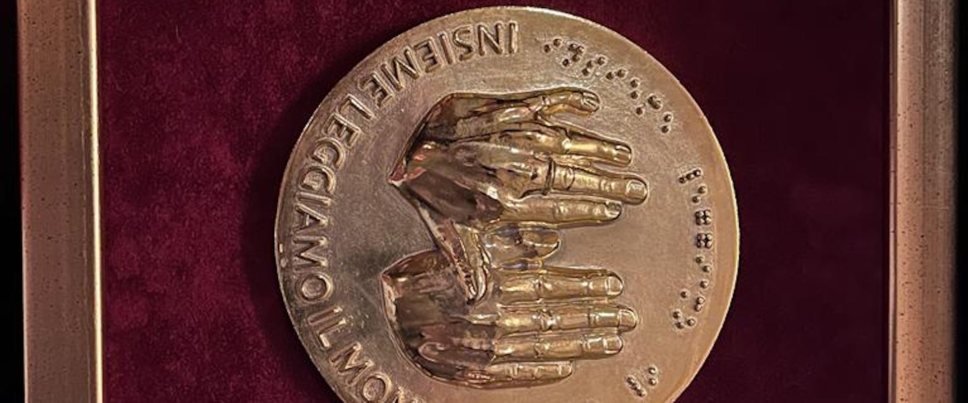 Una medaglia in bronzo con un rilievo di mani che comunicano tramite la lingua dei segni, incorniciata su sfondo rosso, celebrante la Federazione Mondiale dei Sordi.