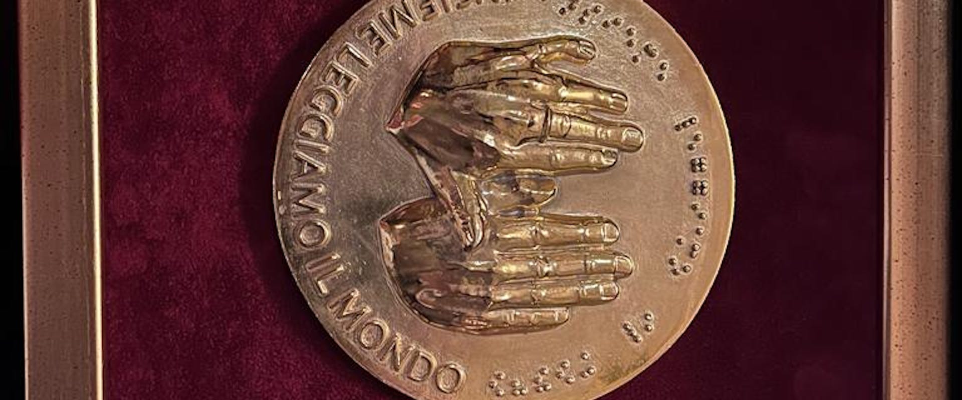 A bronze medal featuring a relief of hands and Braille text, mounted on a burgundy velvet background within a gold frame.