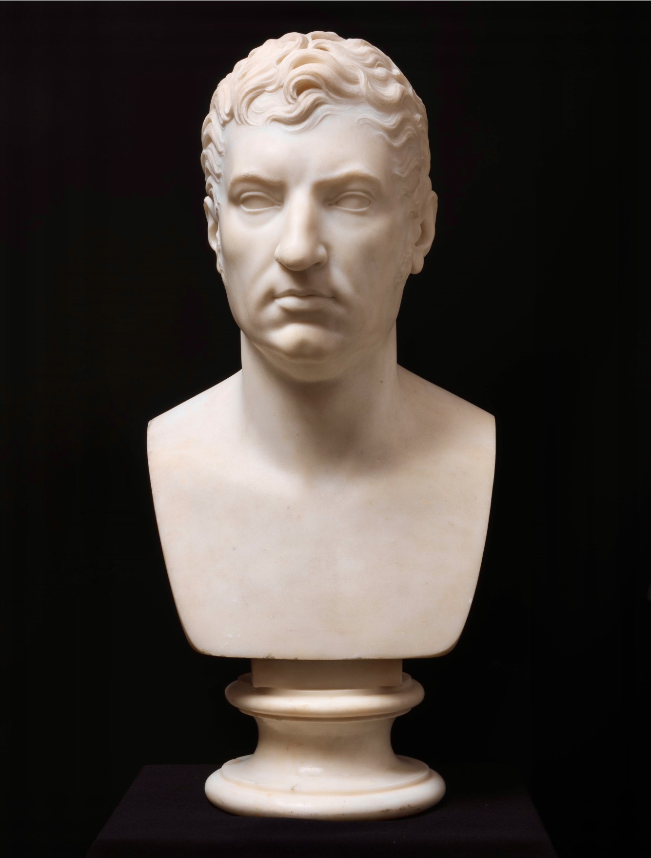 Un busto di marmo bianco di un uomo su uno sfondo nero, che mette in evidenza dettagli accurati dell'acconciatura ondulata e l'espressione seria del volto.