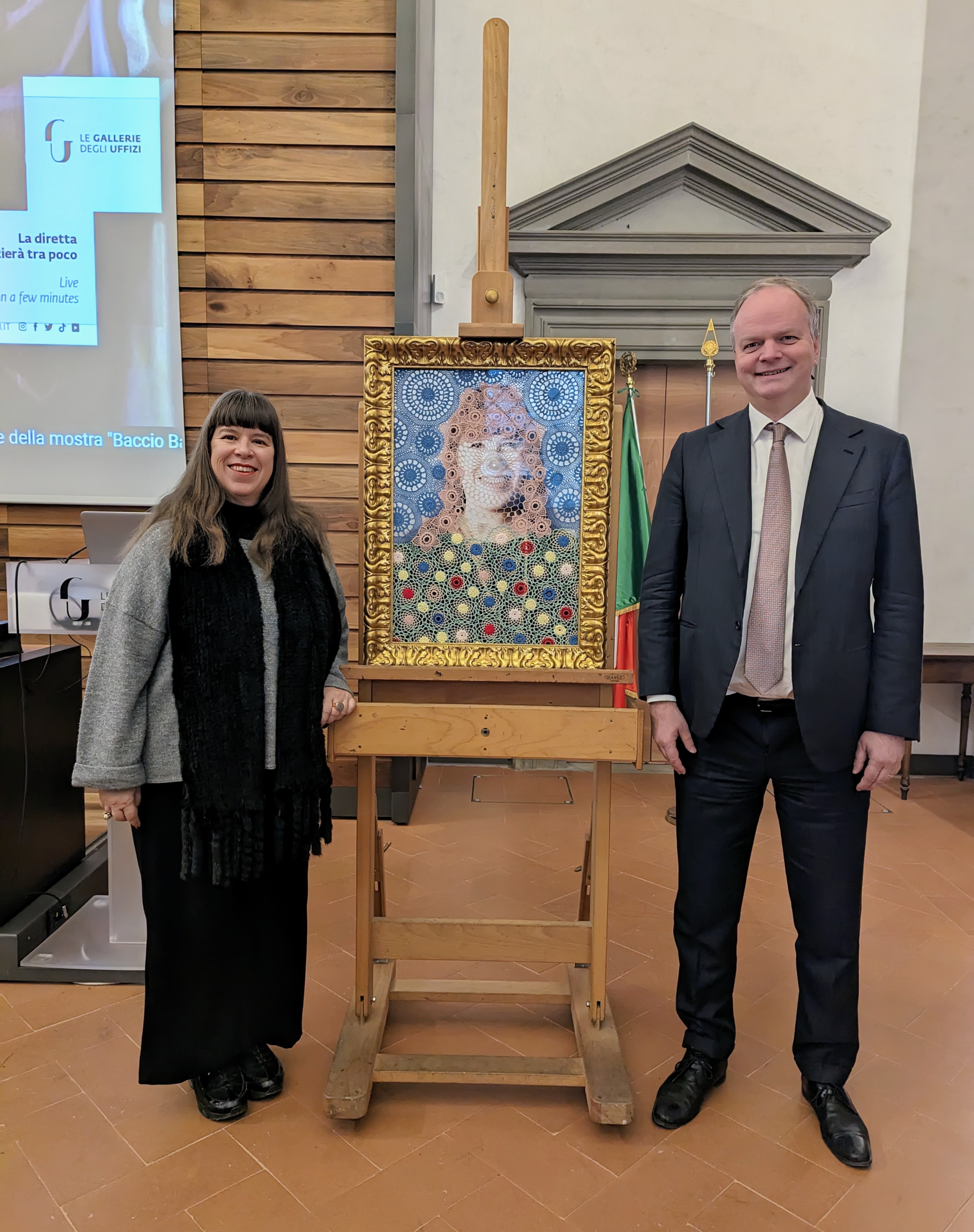 Una donna e un uomo in abito formale posano sorridenti accanto a un cavalletto che sostiene un dipinto riccamente incorniciato, con motivi a cerchi colorati; sullo sfondo, pareti con pannelli di legno e una bandiera italiana visibile.